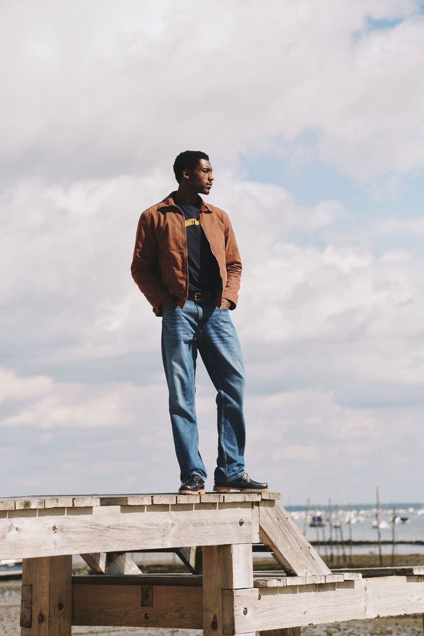 Uomo in piedi su una struttura in legno che indossa una giacca Gant, una maglietta grafica e jeans.