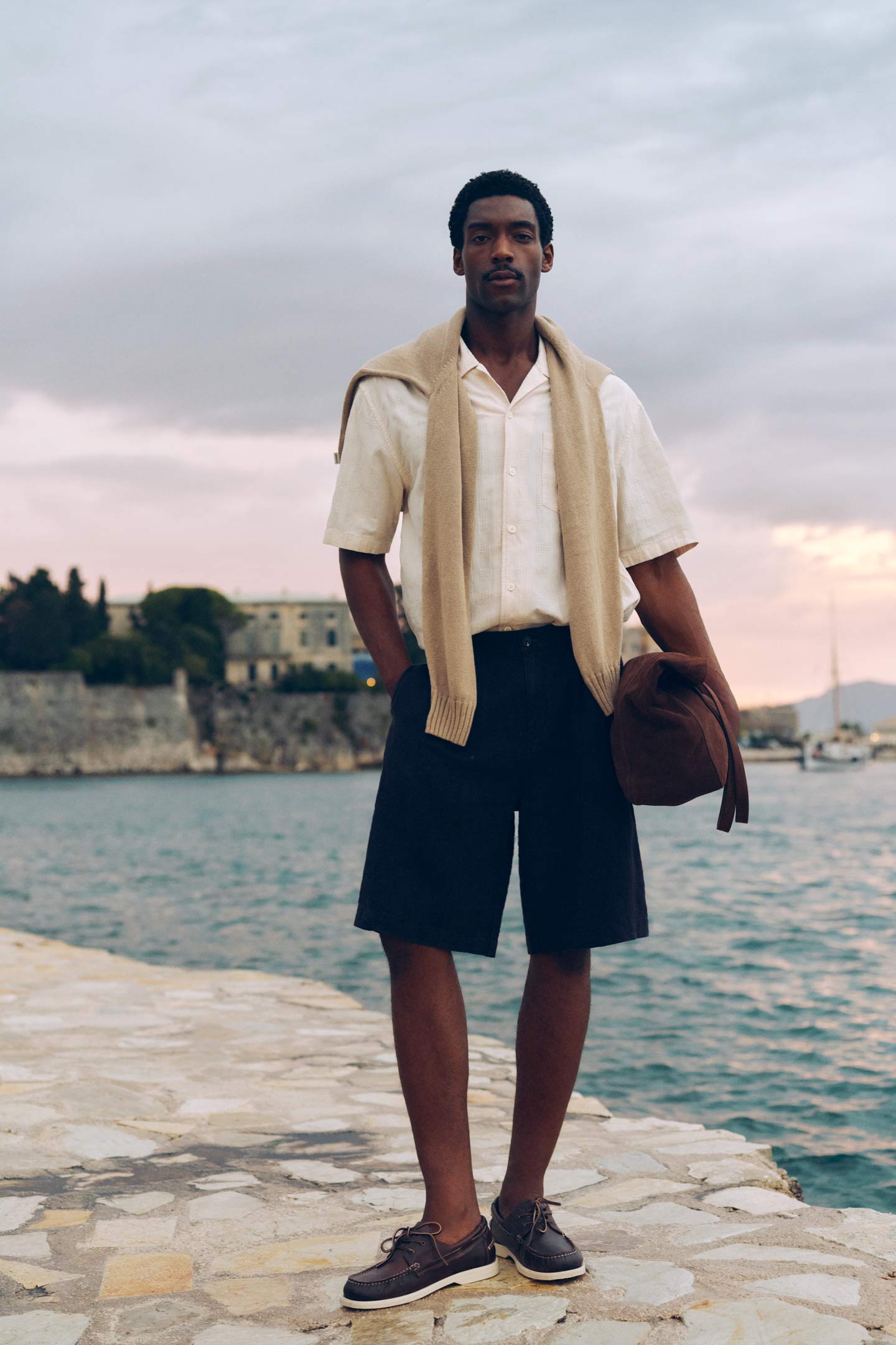Man wearing a Gant linen shirt and shorts standing by the water at sunset.