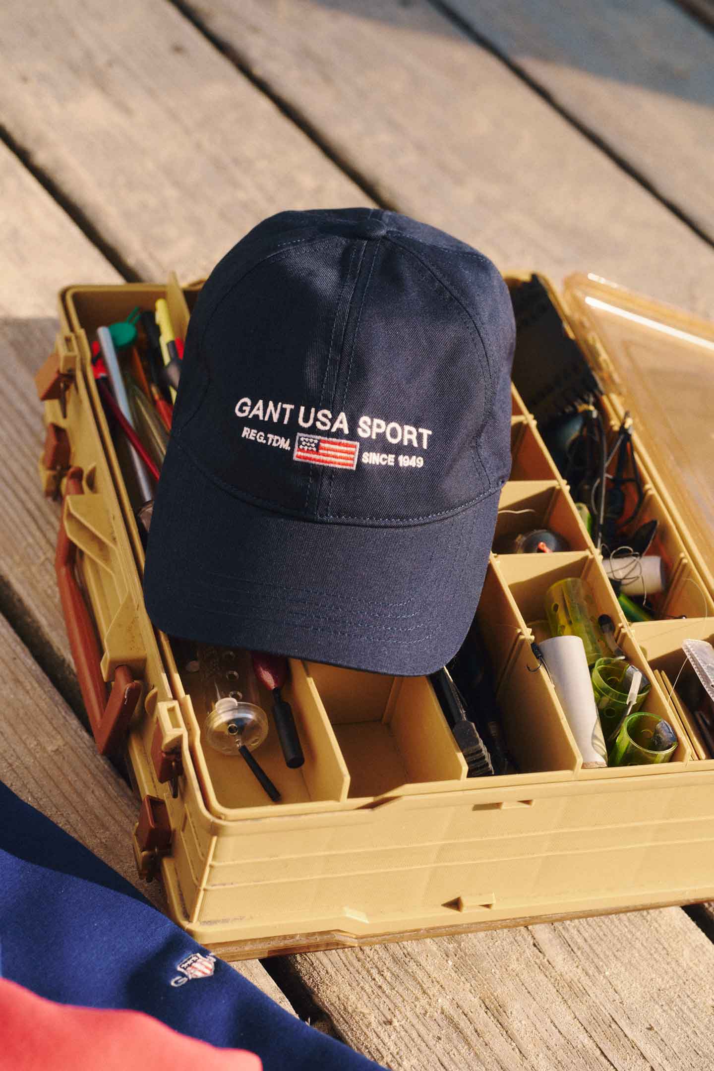 Gant navy sport cap placed on an open tackle box filled with fishing gear on a wooden deck.