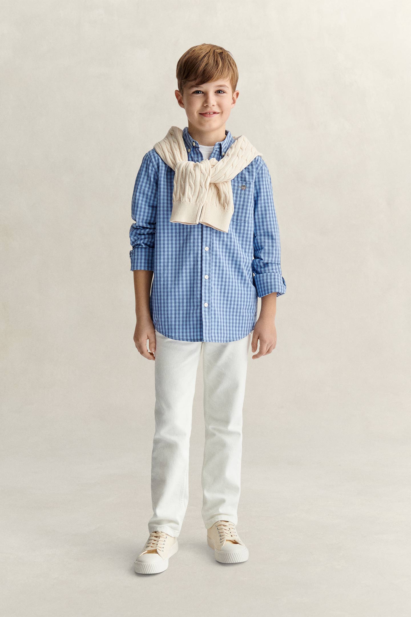 Model styled in a GANT light blue shirt, white pants and cream knitwear over shoulders against a grey backdrop.