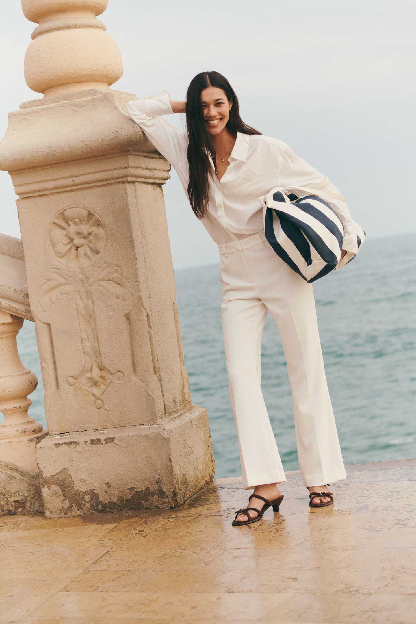 Woman wearing a Gant white shirt and white trousers, holding a canvas striped beach bag by the sea.