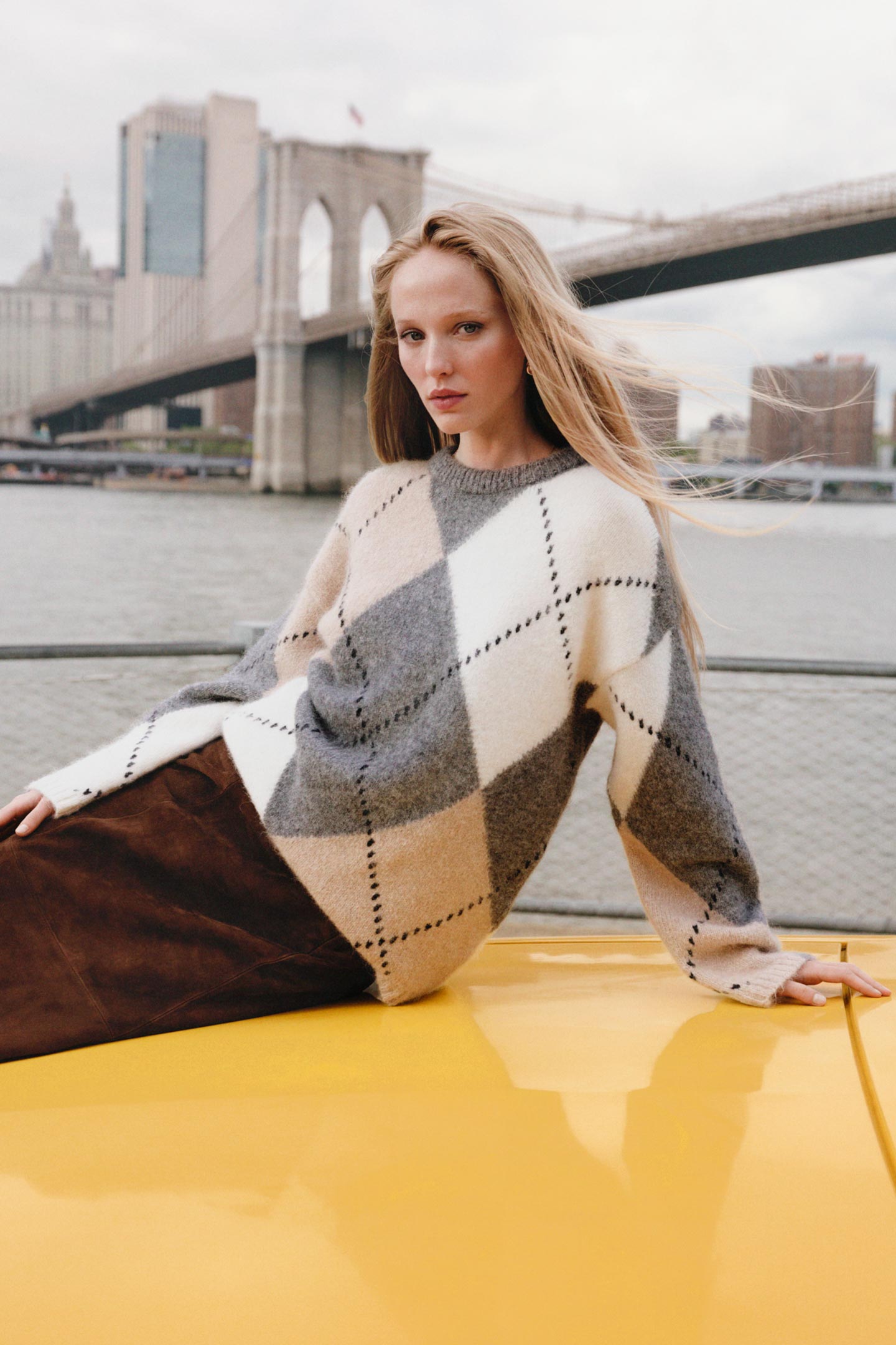 Model styled in a Gant argyle knit sweater paired with a suede skirt, reclining on a yellow taxi near the waterfront.