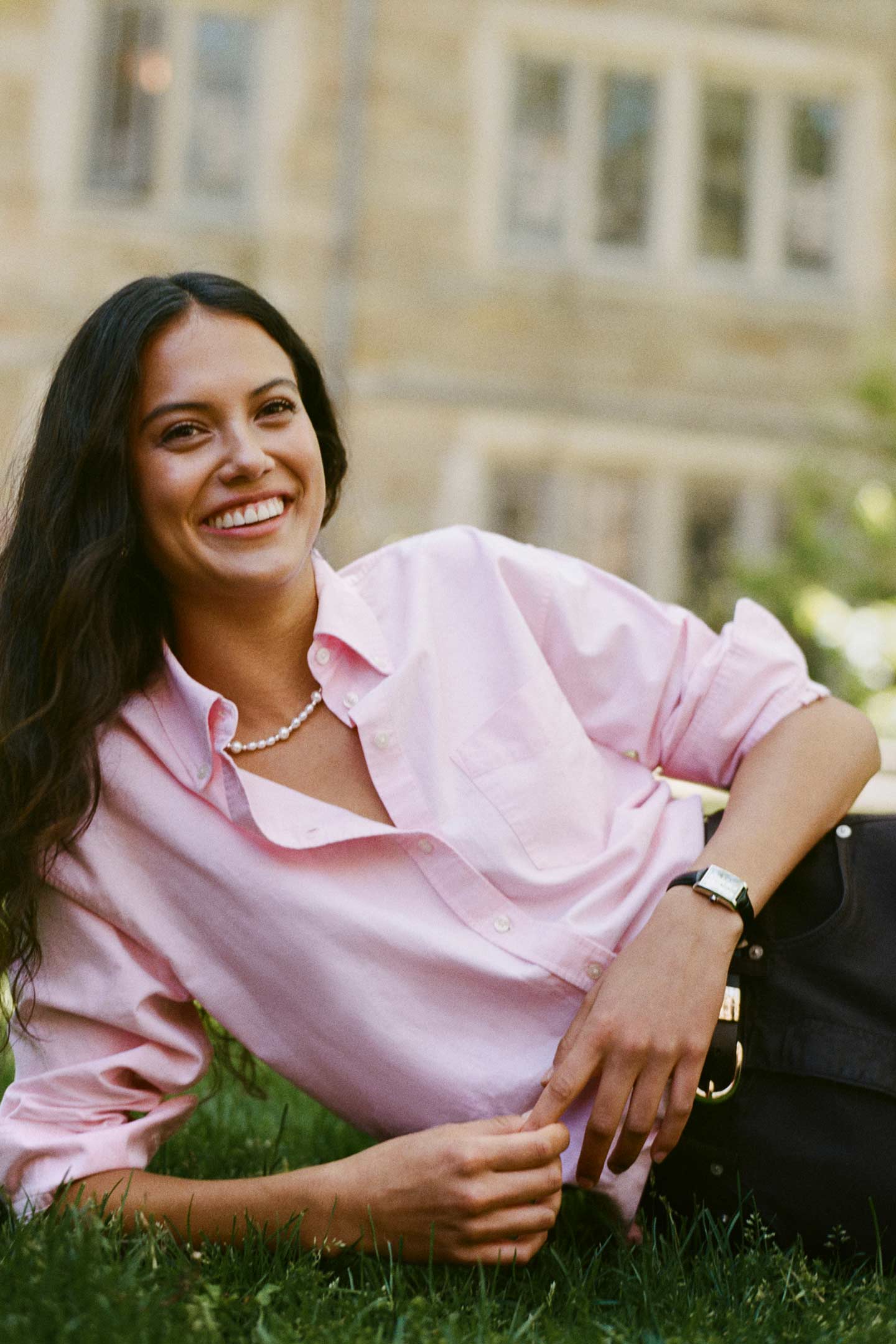 Model styled in a GANT pink shirt with dark jeans lying on grass in an outdoor campus setting.