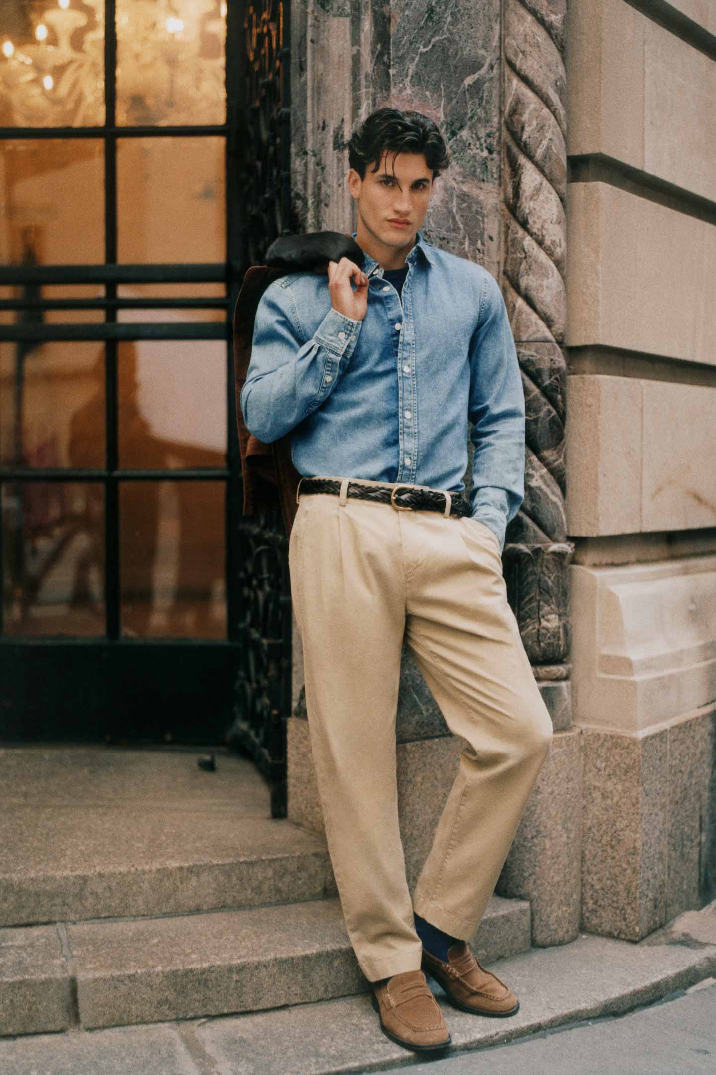 Person wearing a blue denim Gant shirt tucked into beige chinos with loafers outside a stone building.