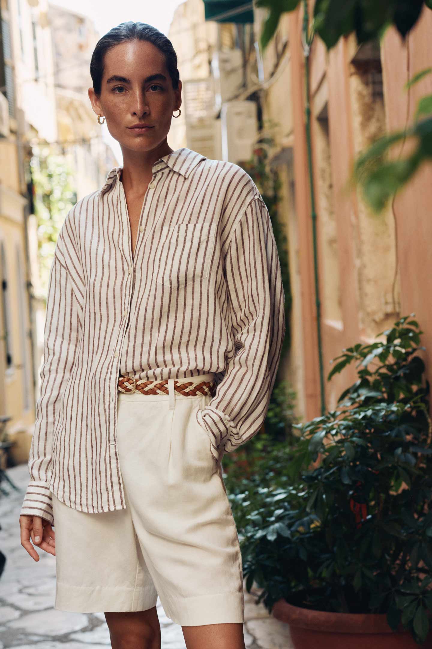 Woman wearing a Gant brown striped button-down shirt and light shorts, standing in a narrow outdoor walkway.
