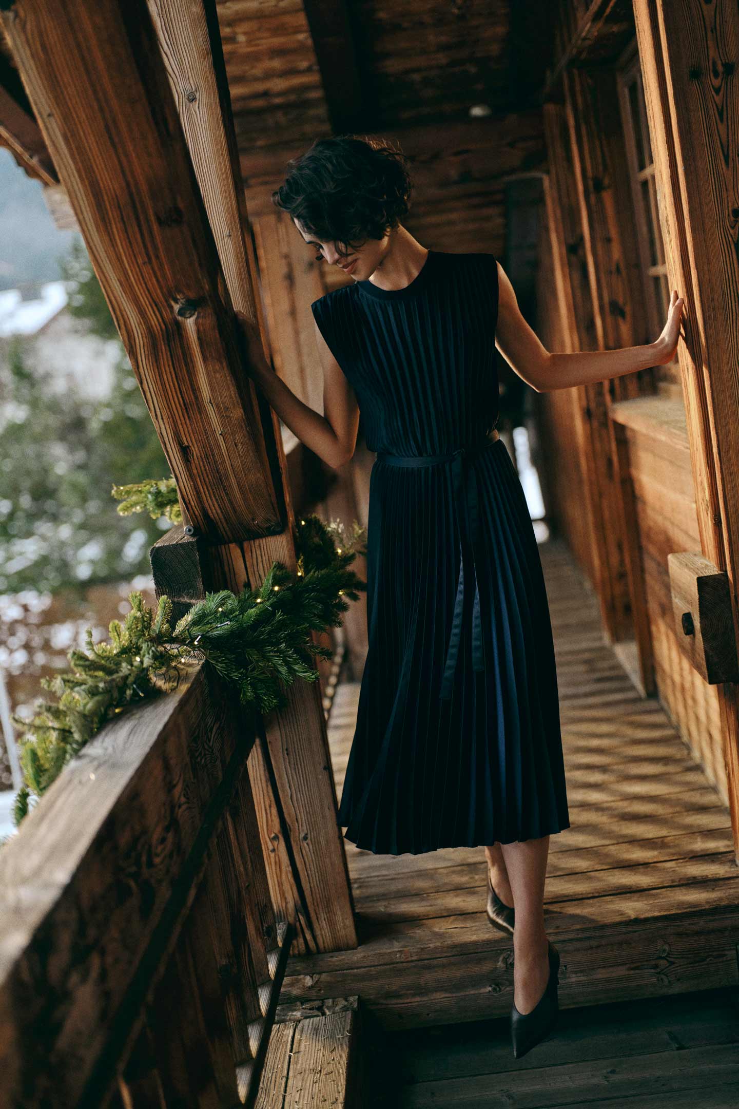 Model styled in a Gant pleated midi dress with a waist belt, standing on a wooden lodge balcony.
