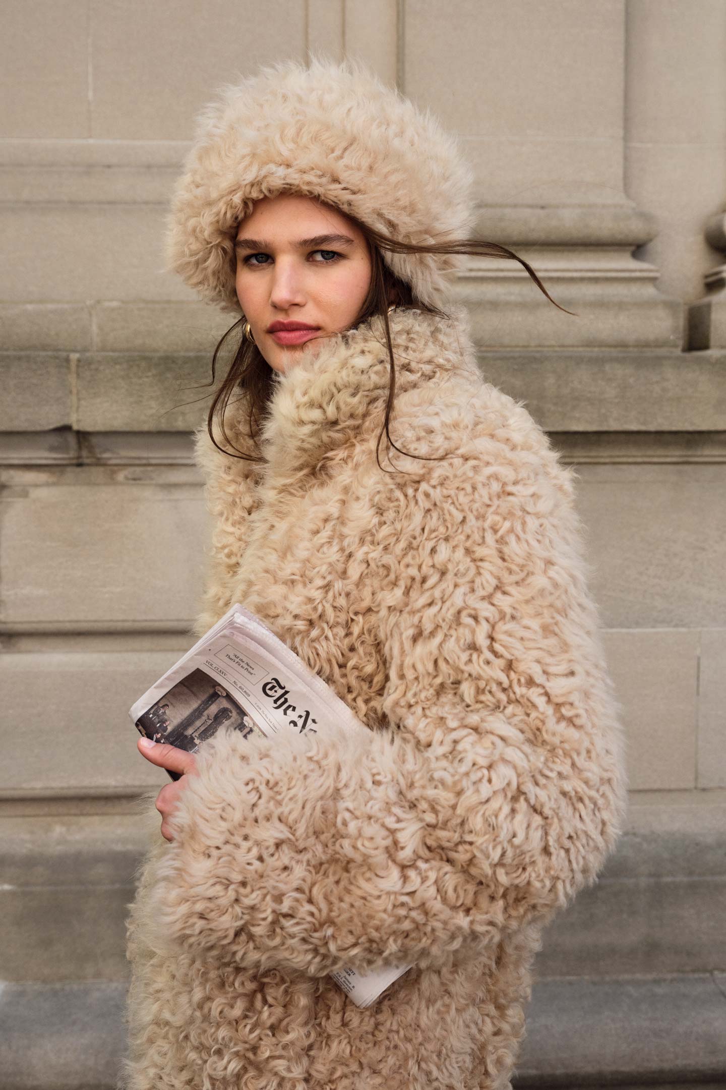 Model styled in a Gant shearling coat and matching hat holding a newspaper under arm.