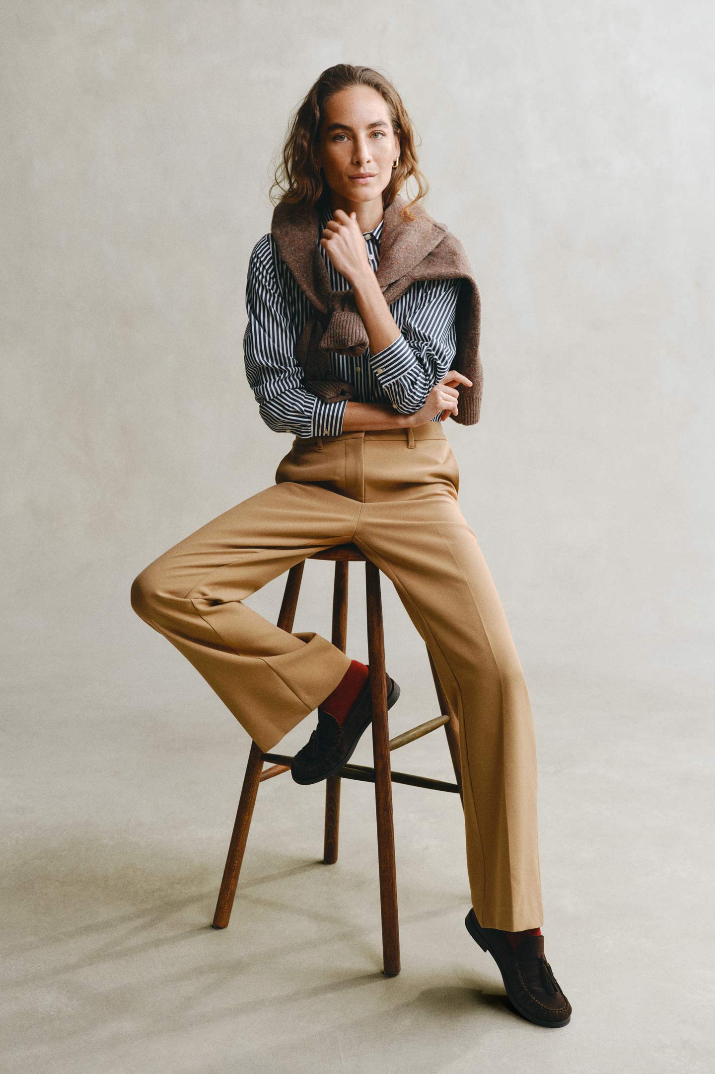 Model styled in a GANT striped shirt paired with beige pants and brown knit over shoulders seated on a stool.