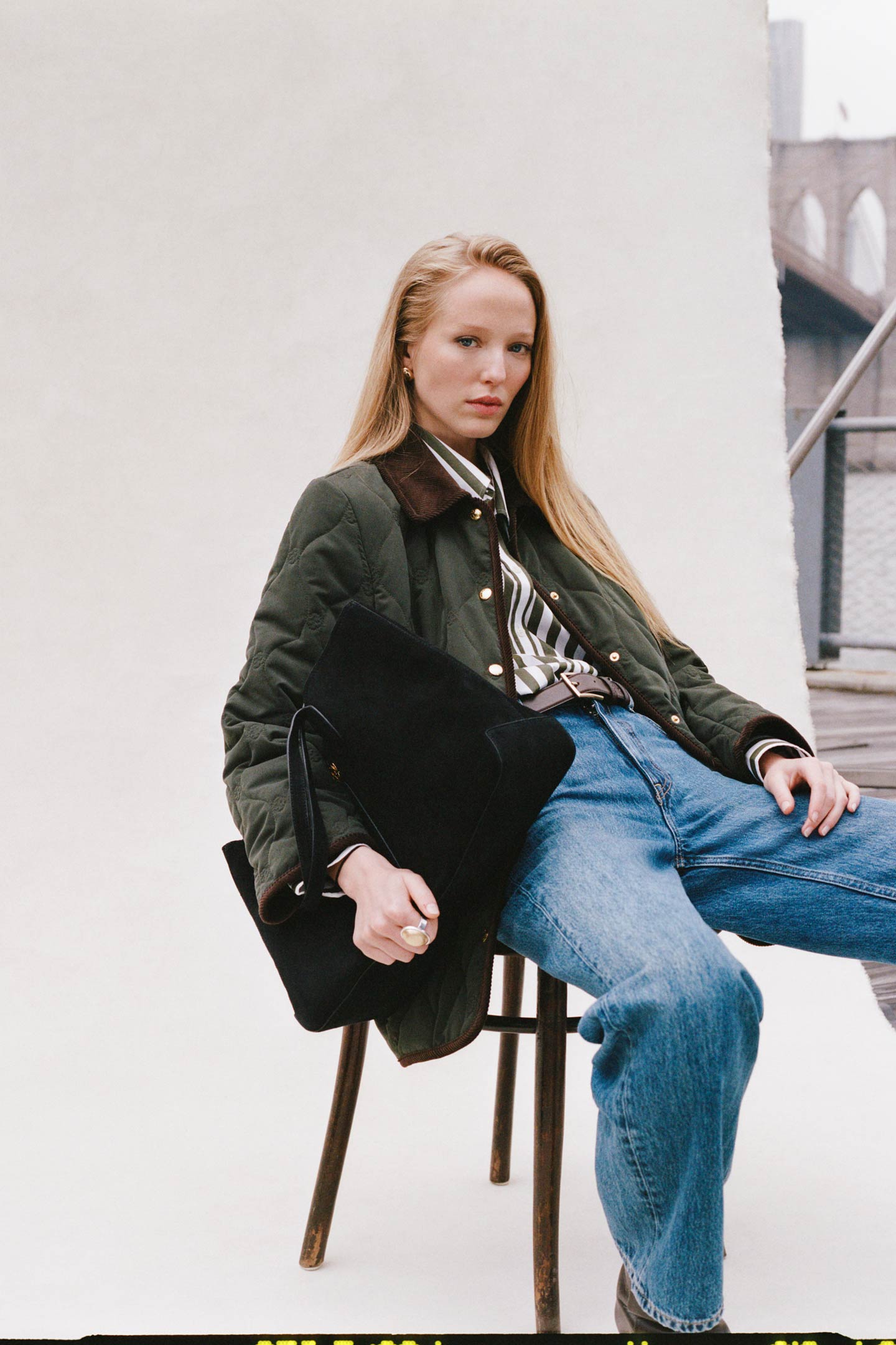 Model styled in a Gant quilted jacket over a striped shirt and jeans, seated on a chair holding a black tote bag.