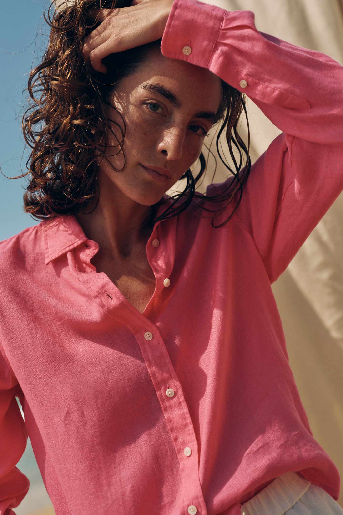Woman wearing a bright pink linen button‑up shirt, standing outdoors with one arm raised toward the sunlight. The background shows a clear blue sky and a light fabric backdrop.