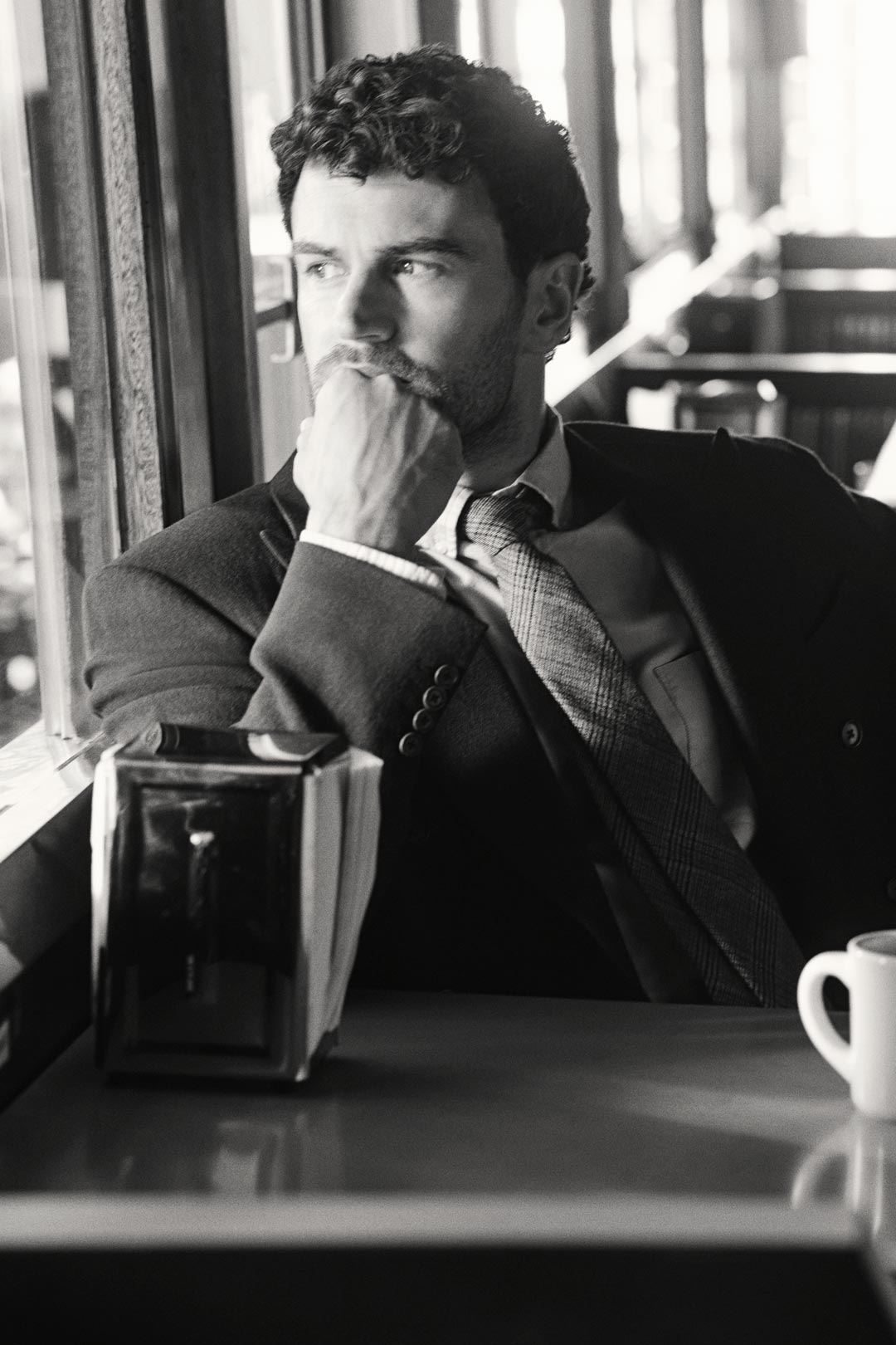 Actor Theo James styled in a GANT tailored blazer with shirt and tie, seated in a diner booth beside a coffee cup.