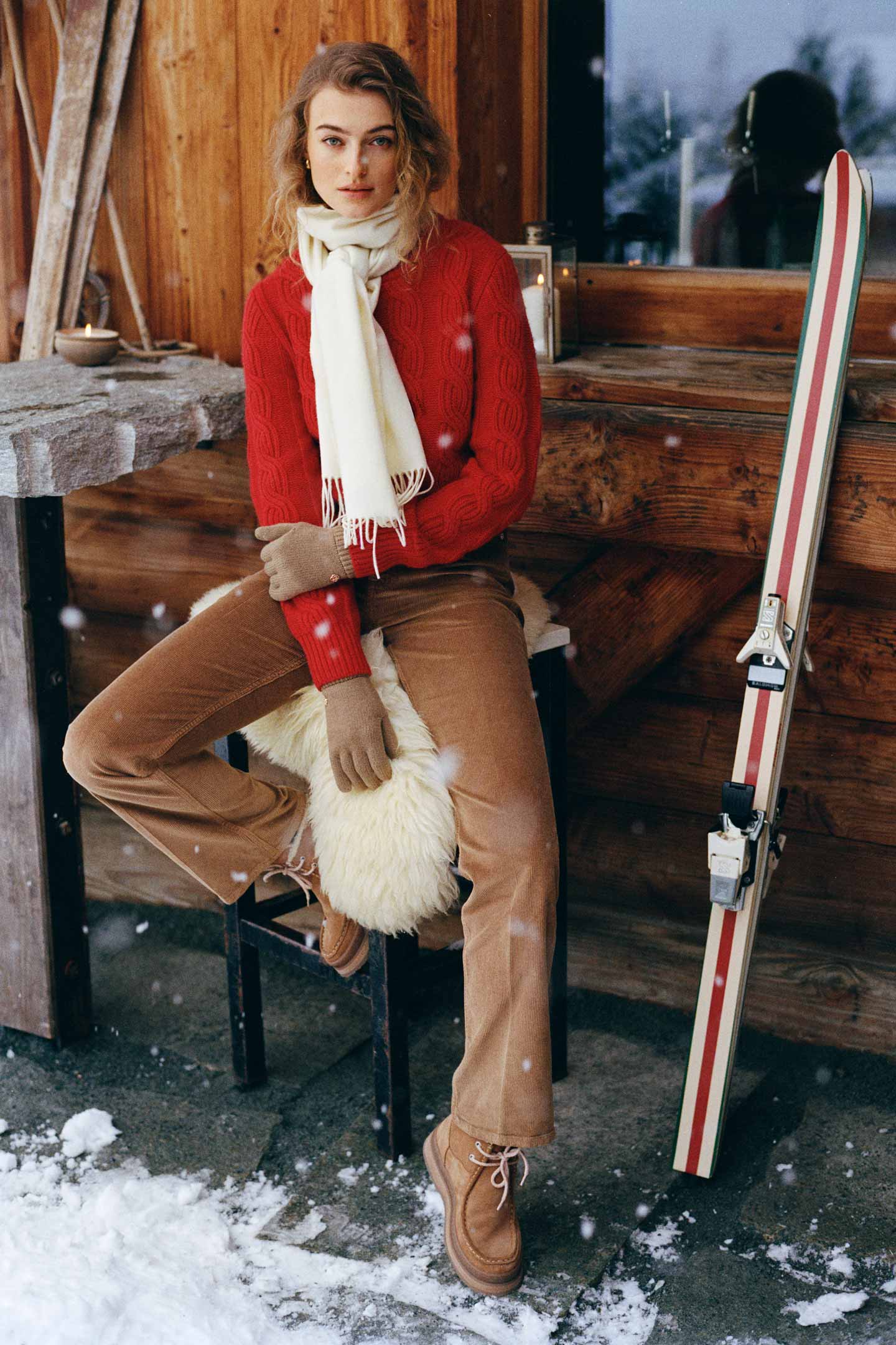 Model styled in a Gant cable-knit sweater paired with a scarf, corduroy pants and boots, seated beside vintage skis.