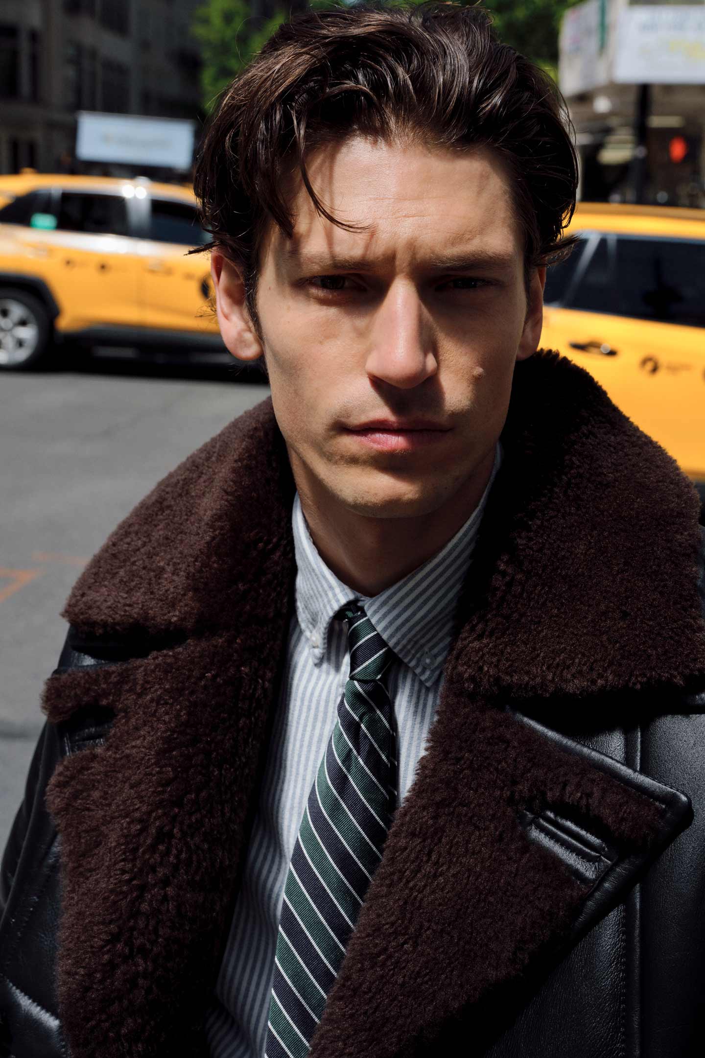 Close up of model styled in a Gant leather coat with shearling trim worn over a shirt and tie, with yellow taxis in the background.