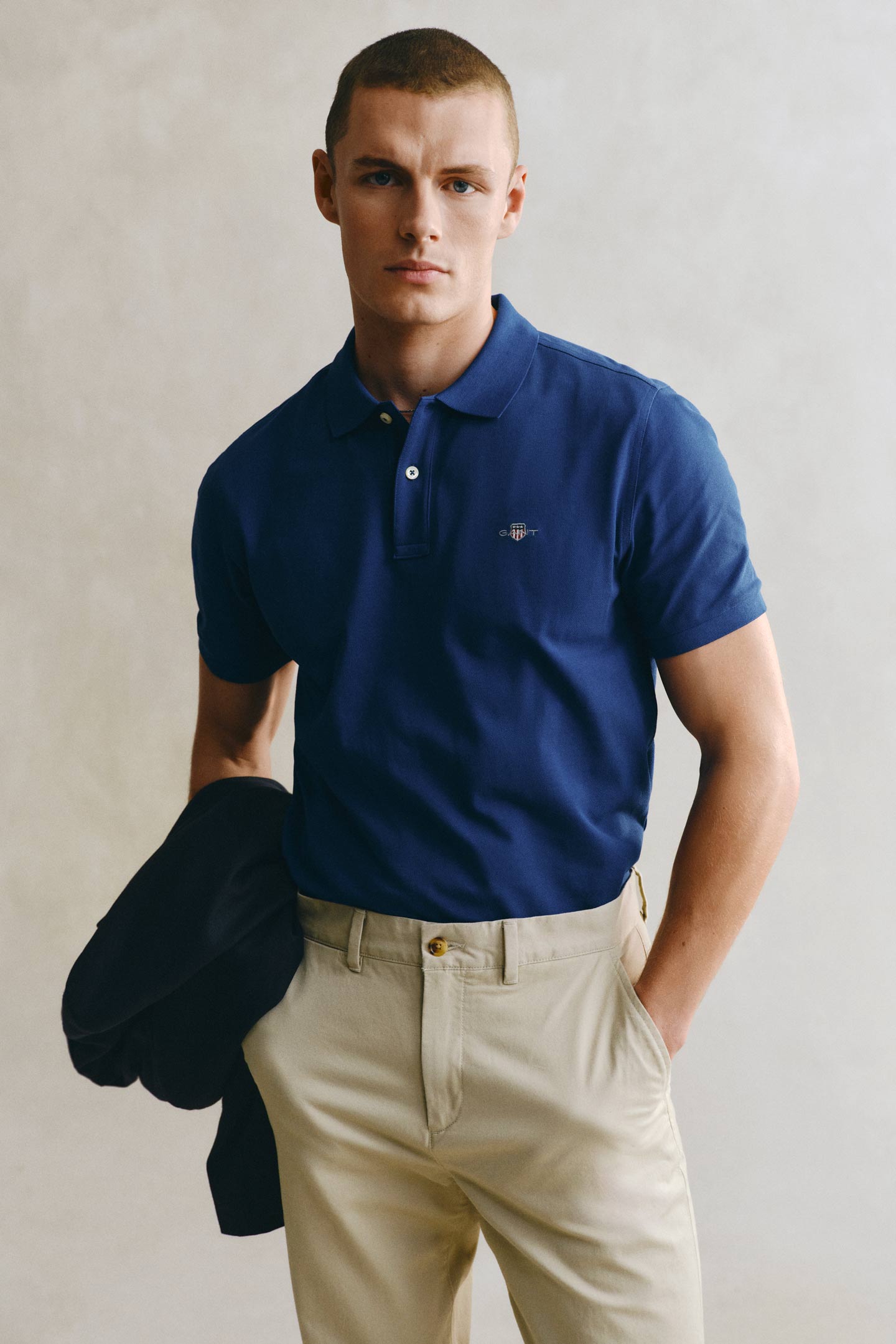 Model styled in a GANT blue polo shirt paired with khaki chinos holding a coat standing in front of a grey backdrop.