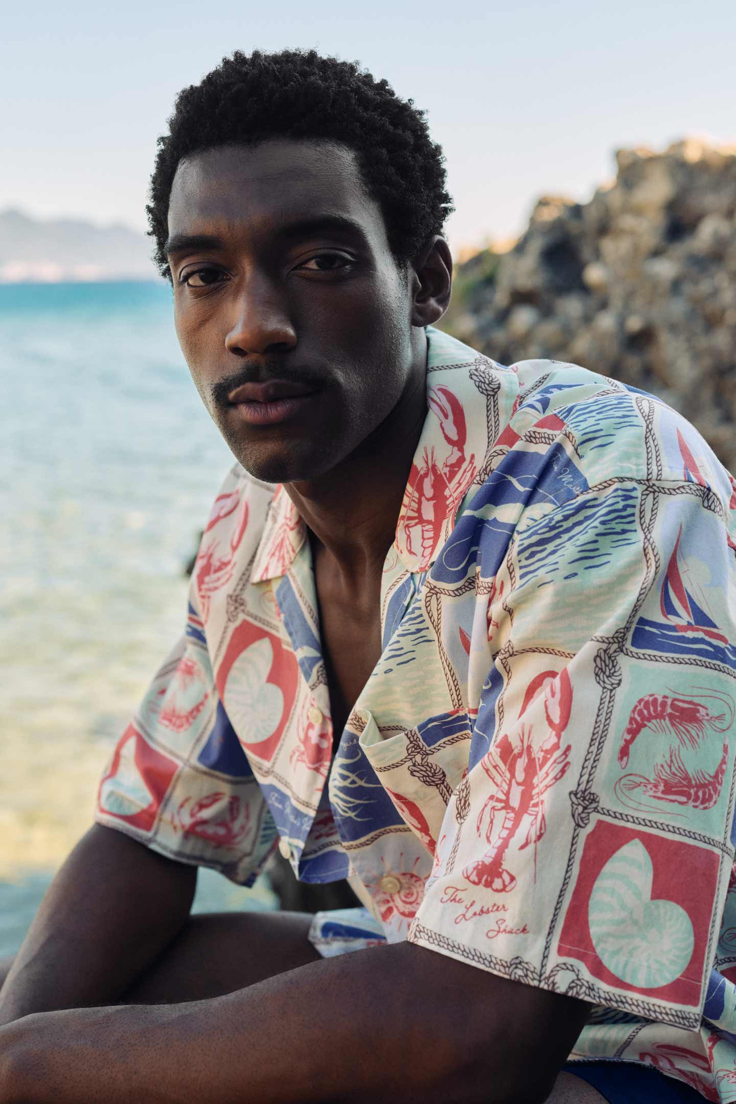 Man wearing a Gant patterned short‑sleeve shirt while sitting near the shoreline.