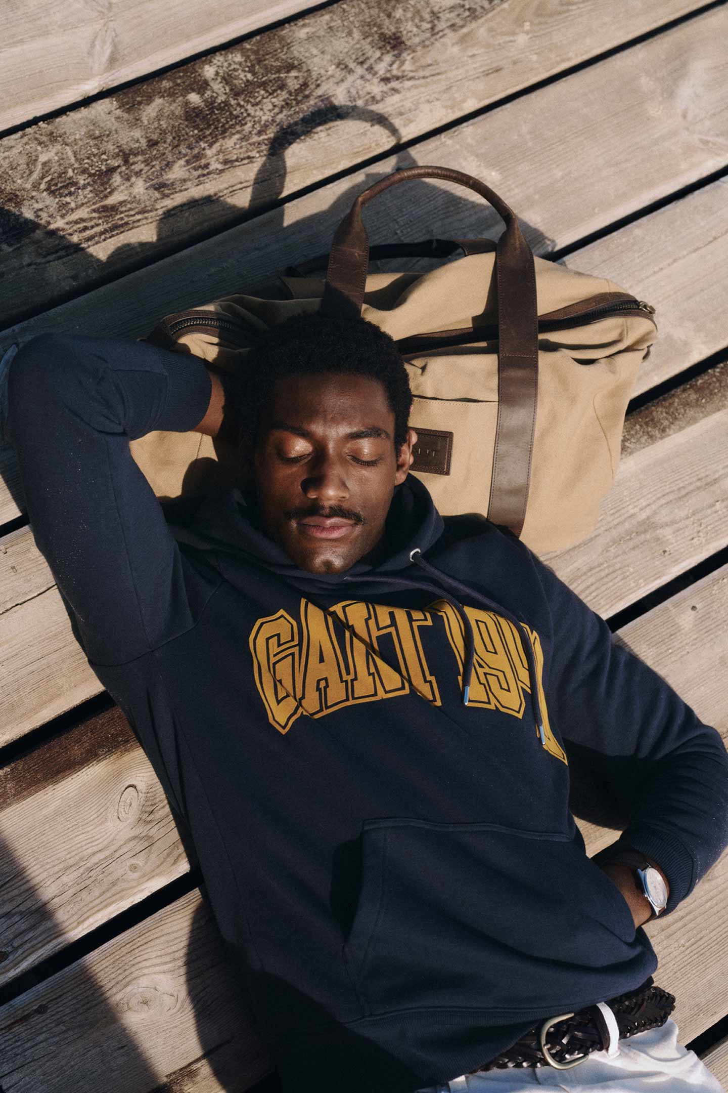 Man liggend op een steiger, gekleed in een marineblauwe Gant-logo hoodie naast een canvas tas.