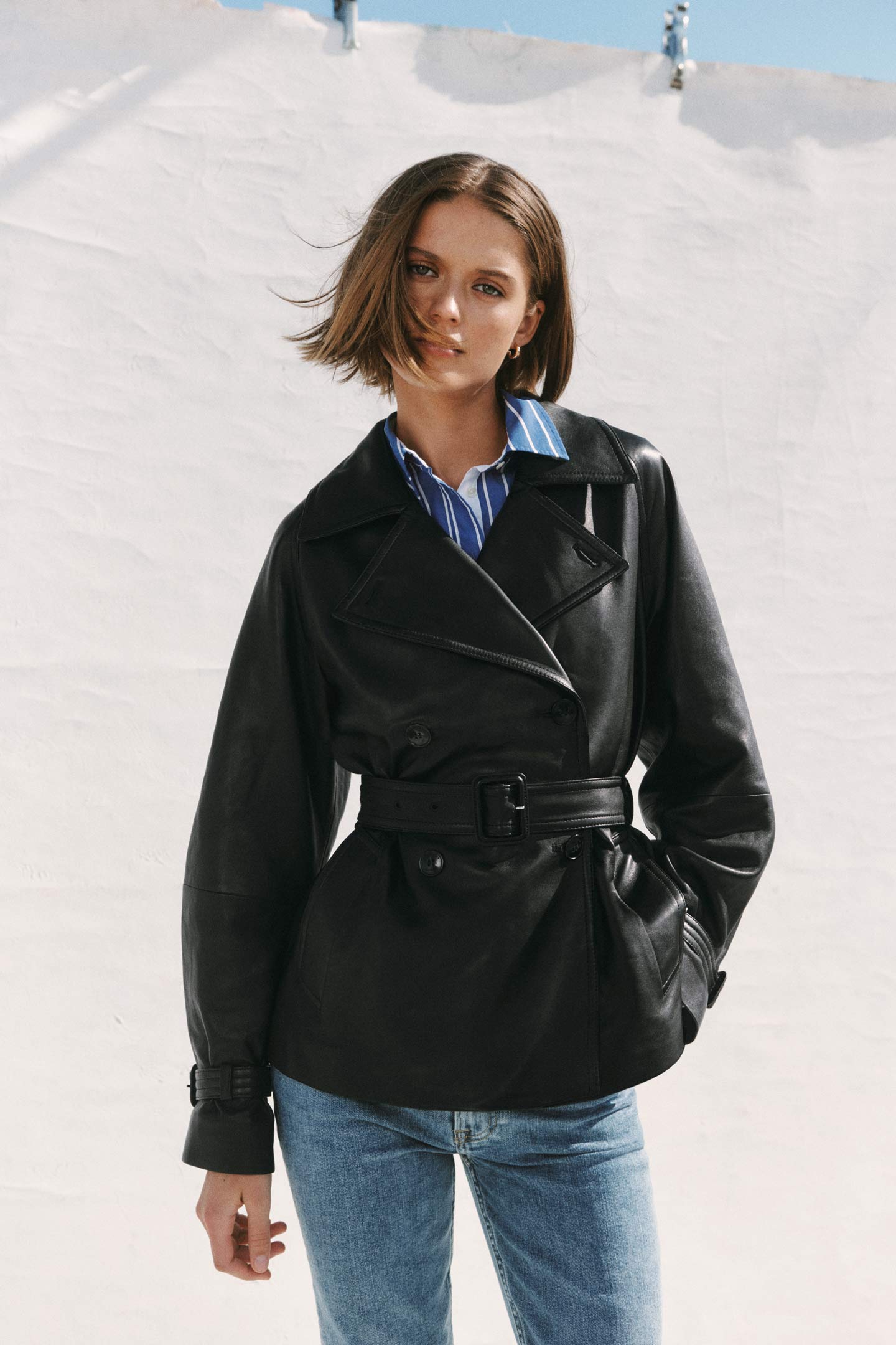 Woman wearing a Gant belted leather jacket over a striped shirt with jeans, standing in an outdoor rooftop setting