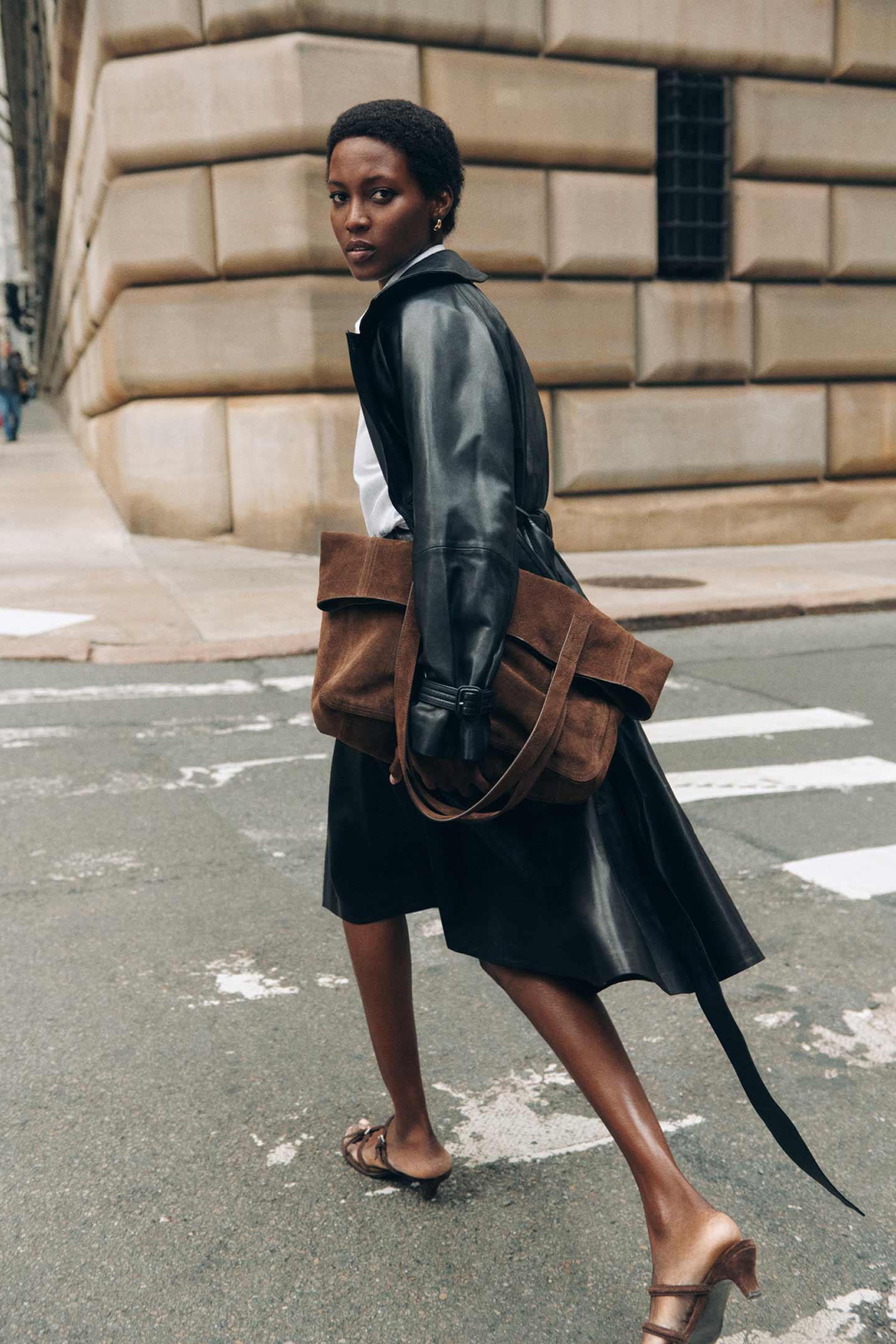 Personne portant un manteau et une jupe en cuir Gant traverse une rue avec un grand sac en daim.