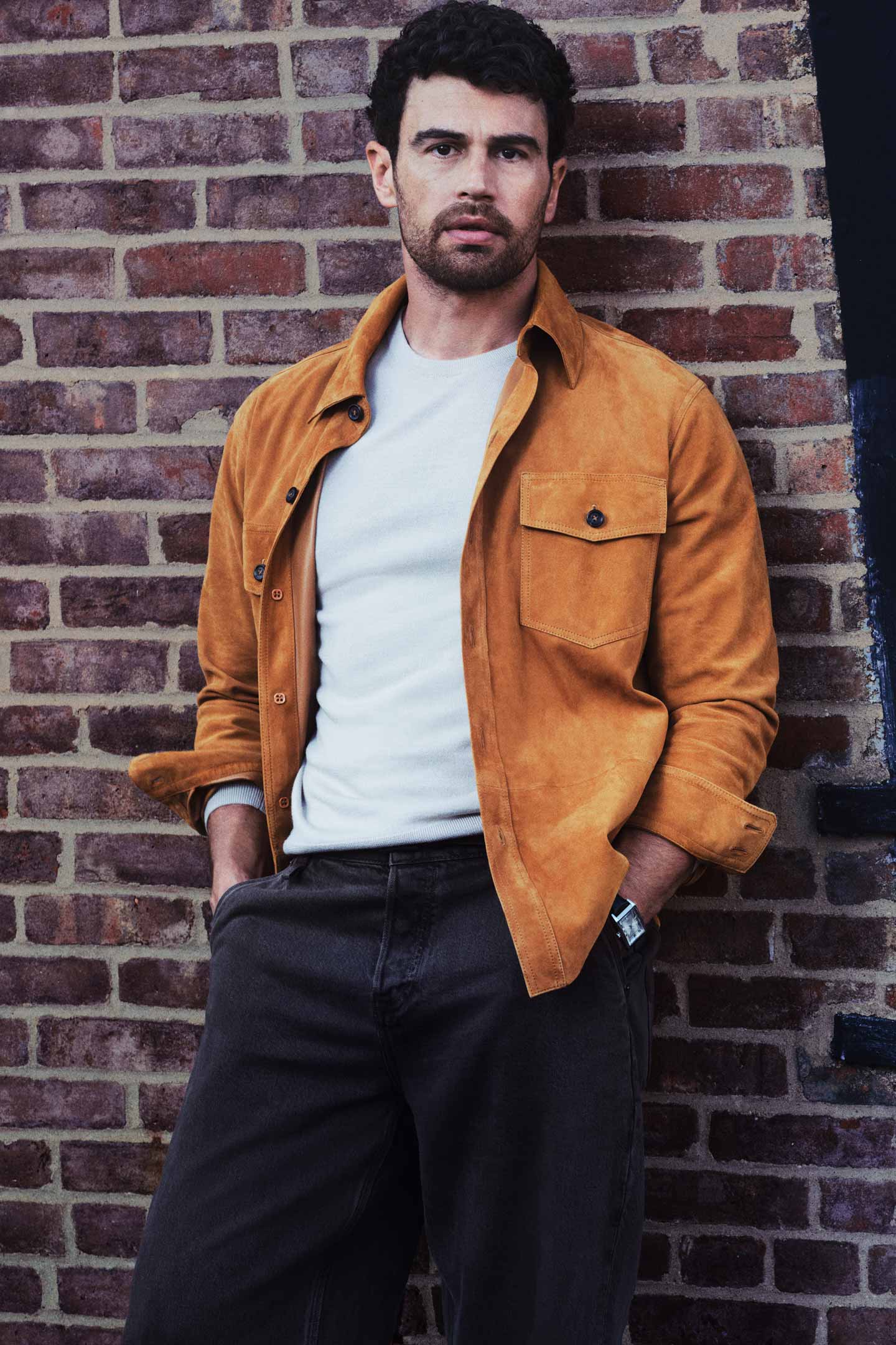 Actor Theo James styled in a GANT suede overshirt worn with a crew neck T-shirt and trousers, standing against a brick wall in an urban setting.