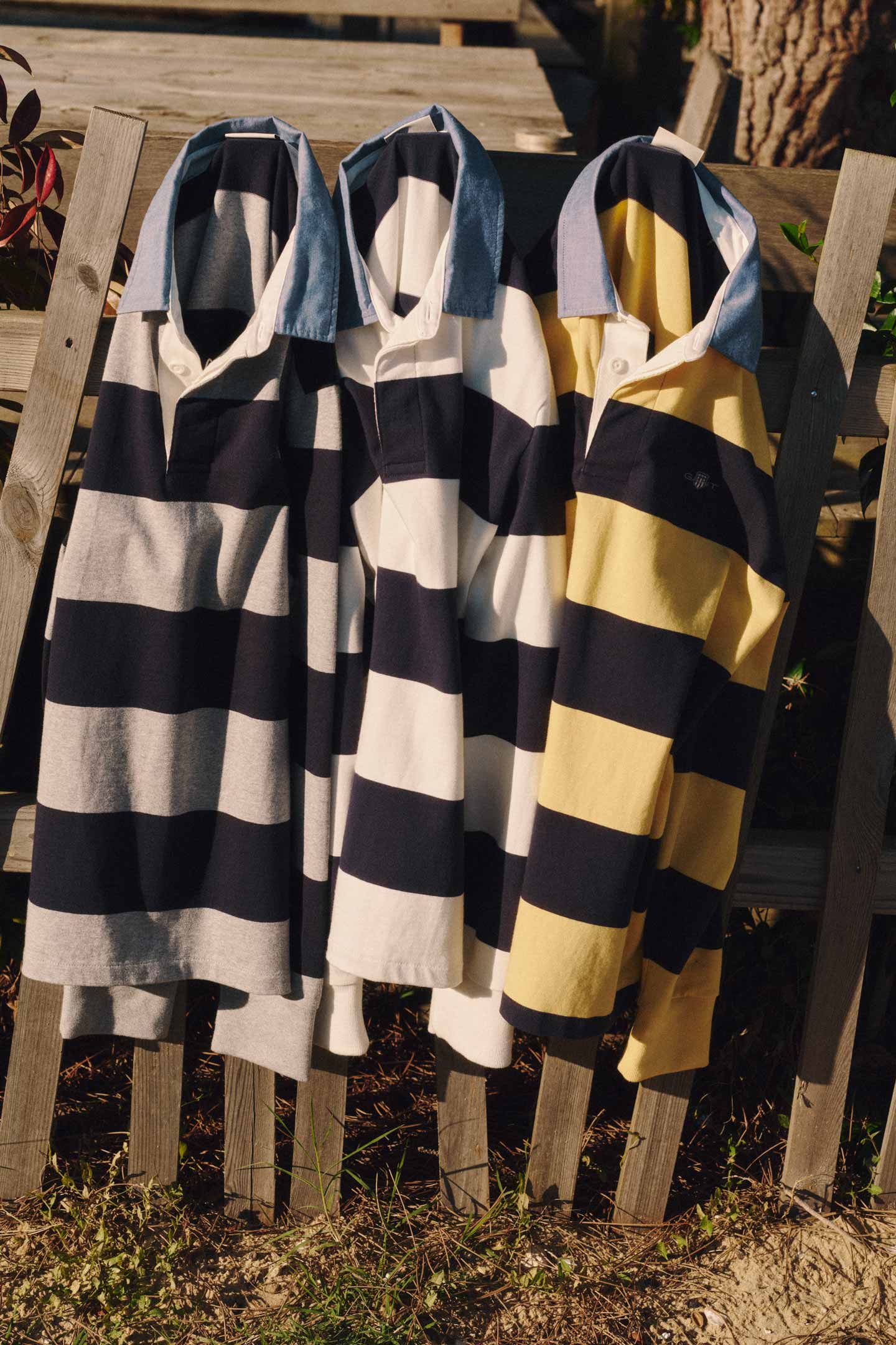 Gant striped rugby shirts in blue‑grey, blue‑white, and blue‑yellow hanging on a wooden fence in sunlight.