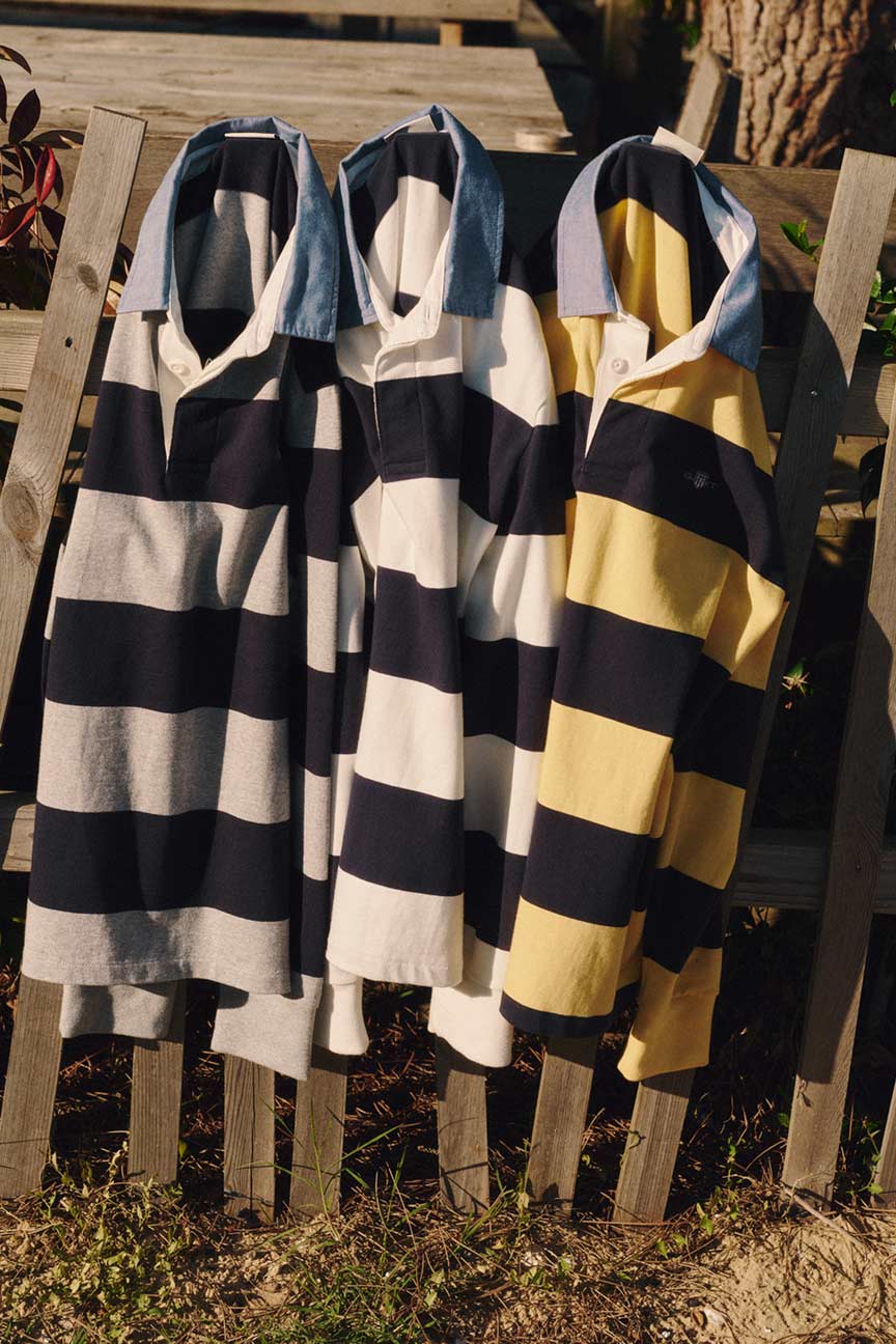 Gant striped rugby shirts in blue‑grey, blue‑white, and blue‑yellow hanging on a wooden fence in sunlight.