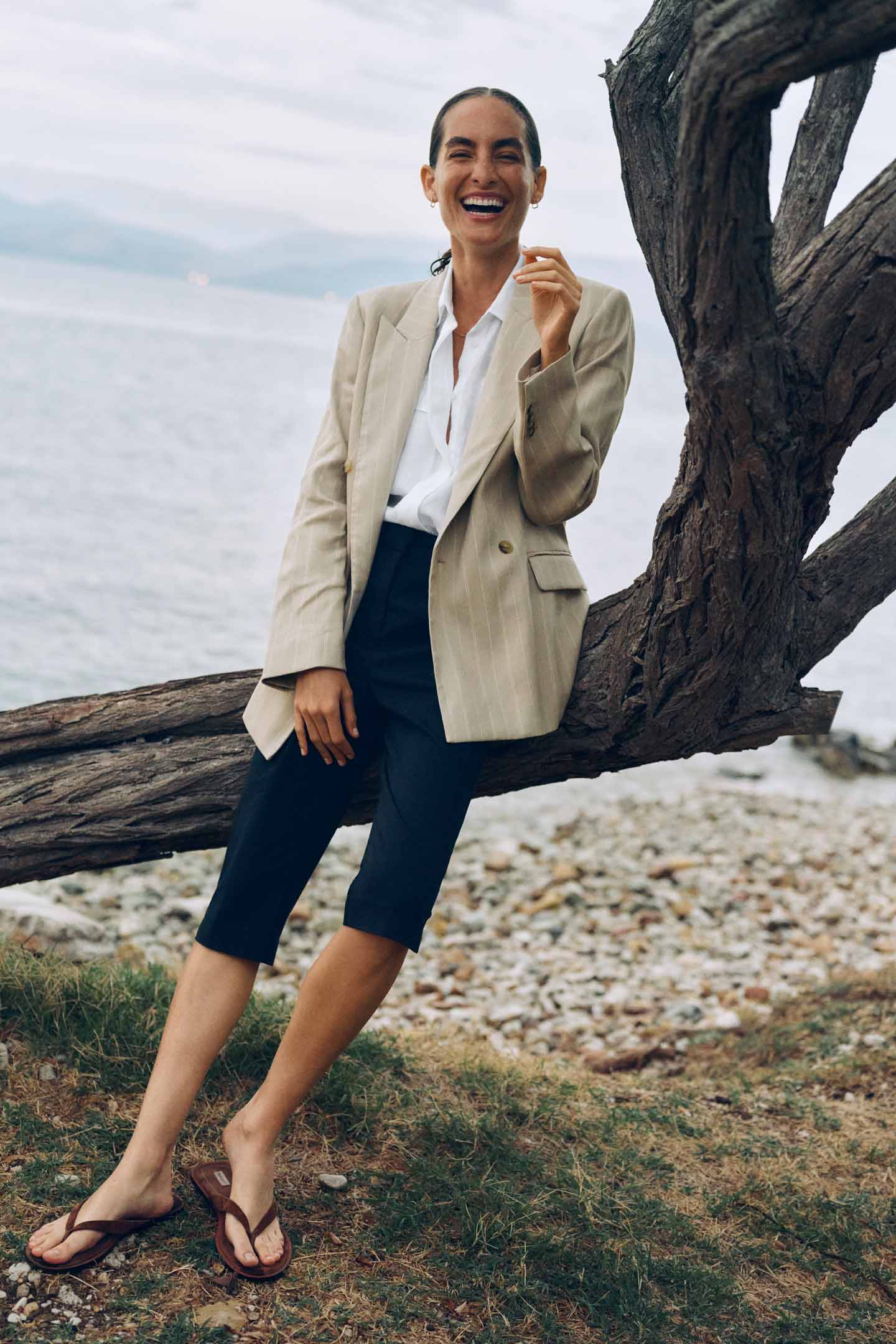 Woman wearing a Gant blazer, white shirt and shorts smiling while leaning against a tree by the water.