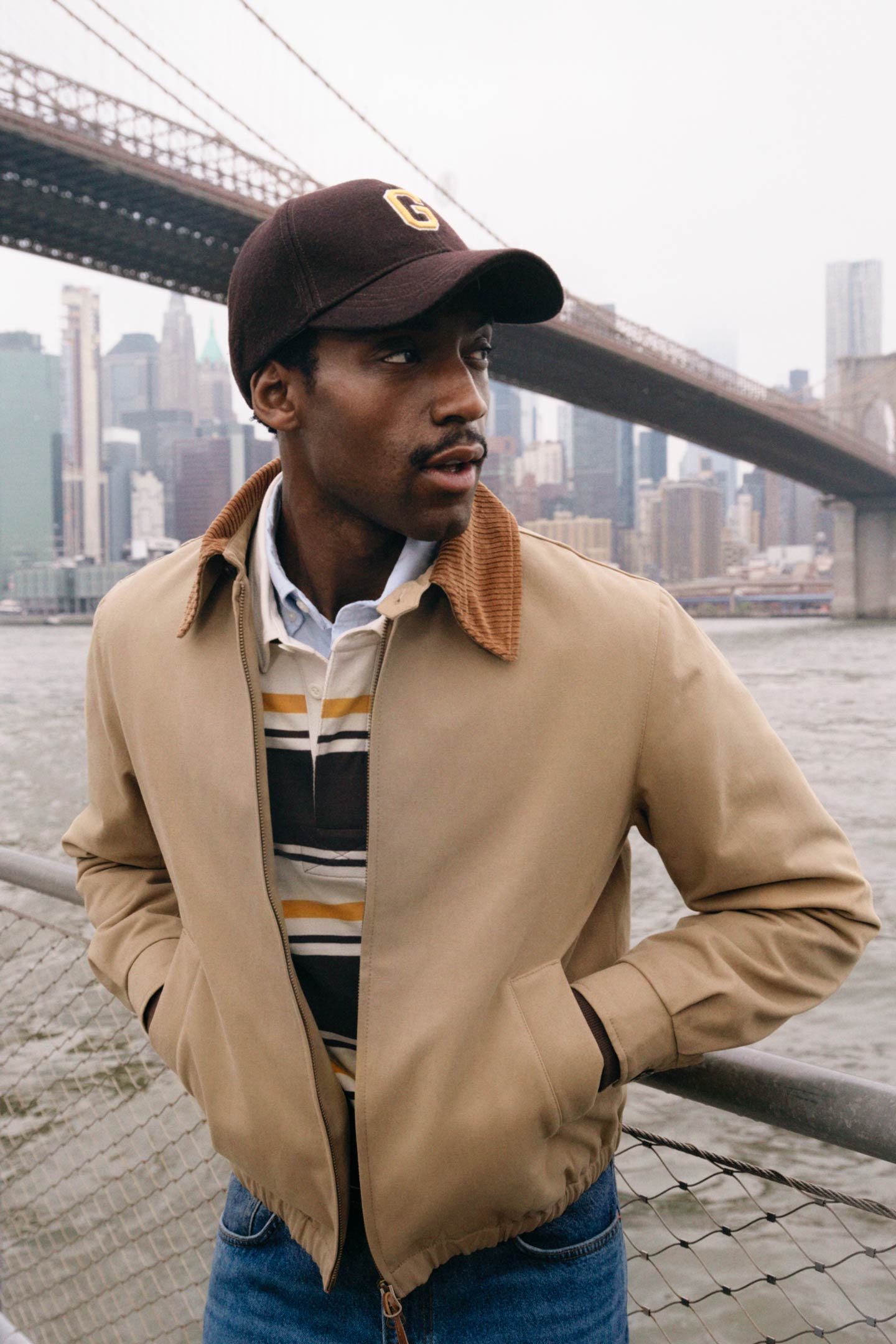 Model styled in a Gant bomber jacket layered over a striped rugby shirt and jeans, standing against railings by the waterfront.