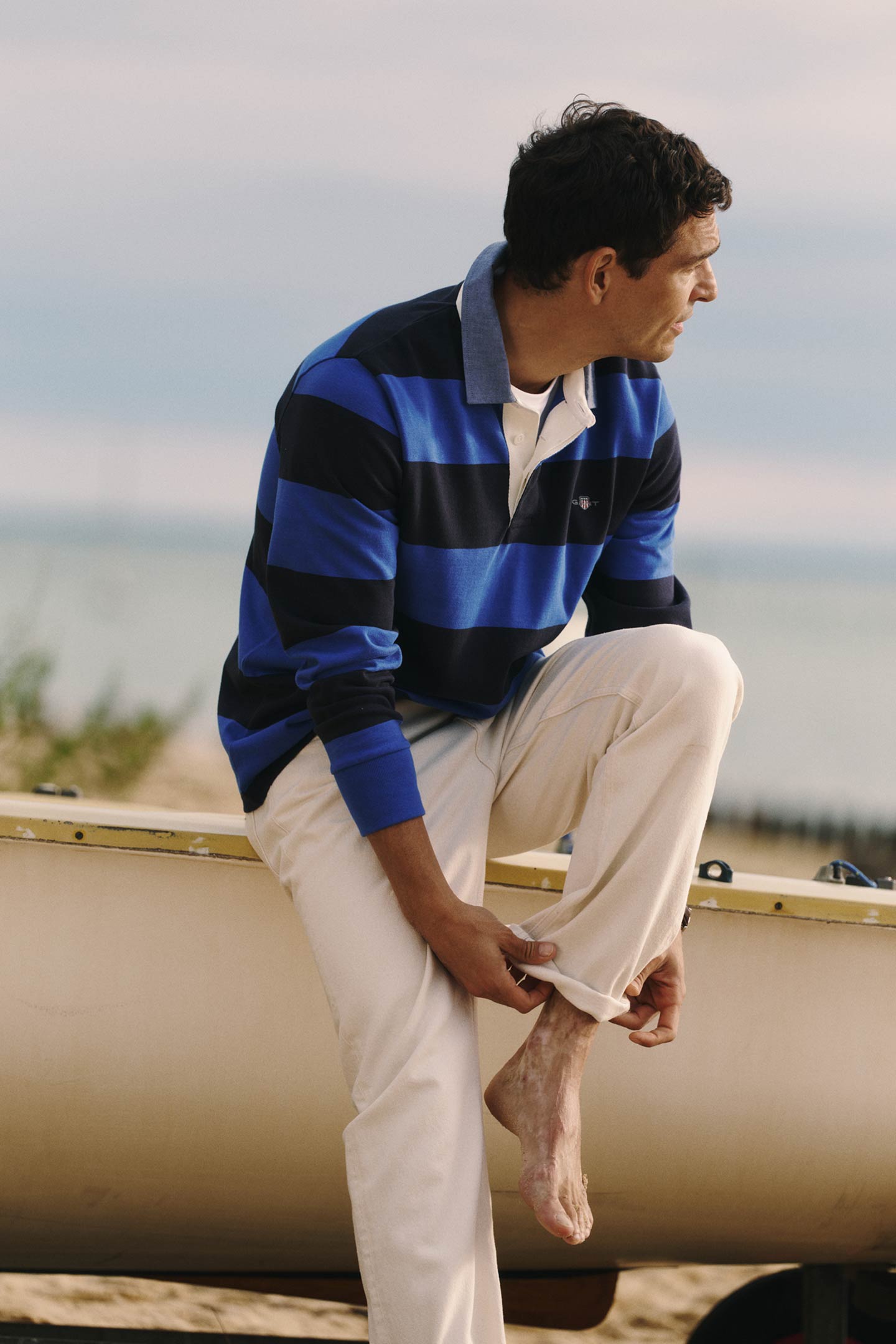 Gant striped rugby shirt in blue and navy worn by a men sitting on a boat and adjusting a pant leg.