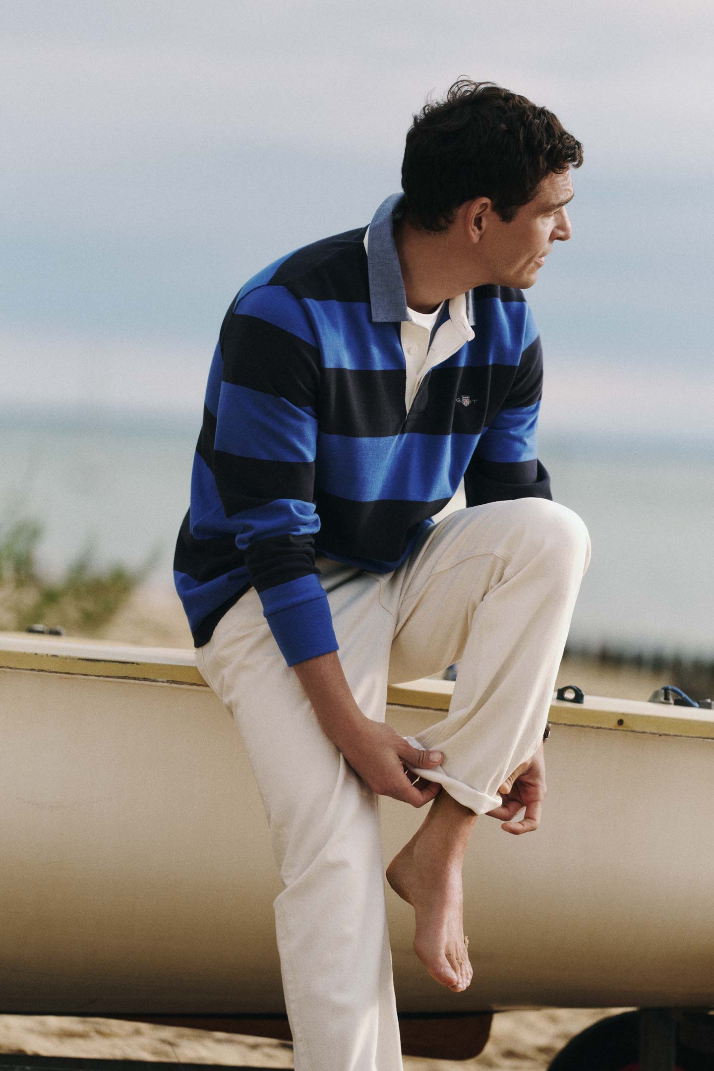 Gant striped rugby shirt in blue and navy worn by a men sitting on a boat and adjusting a pant leg.