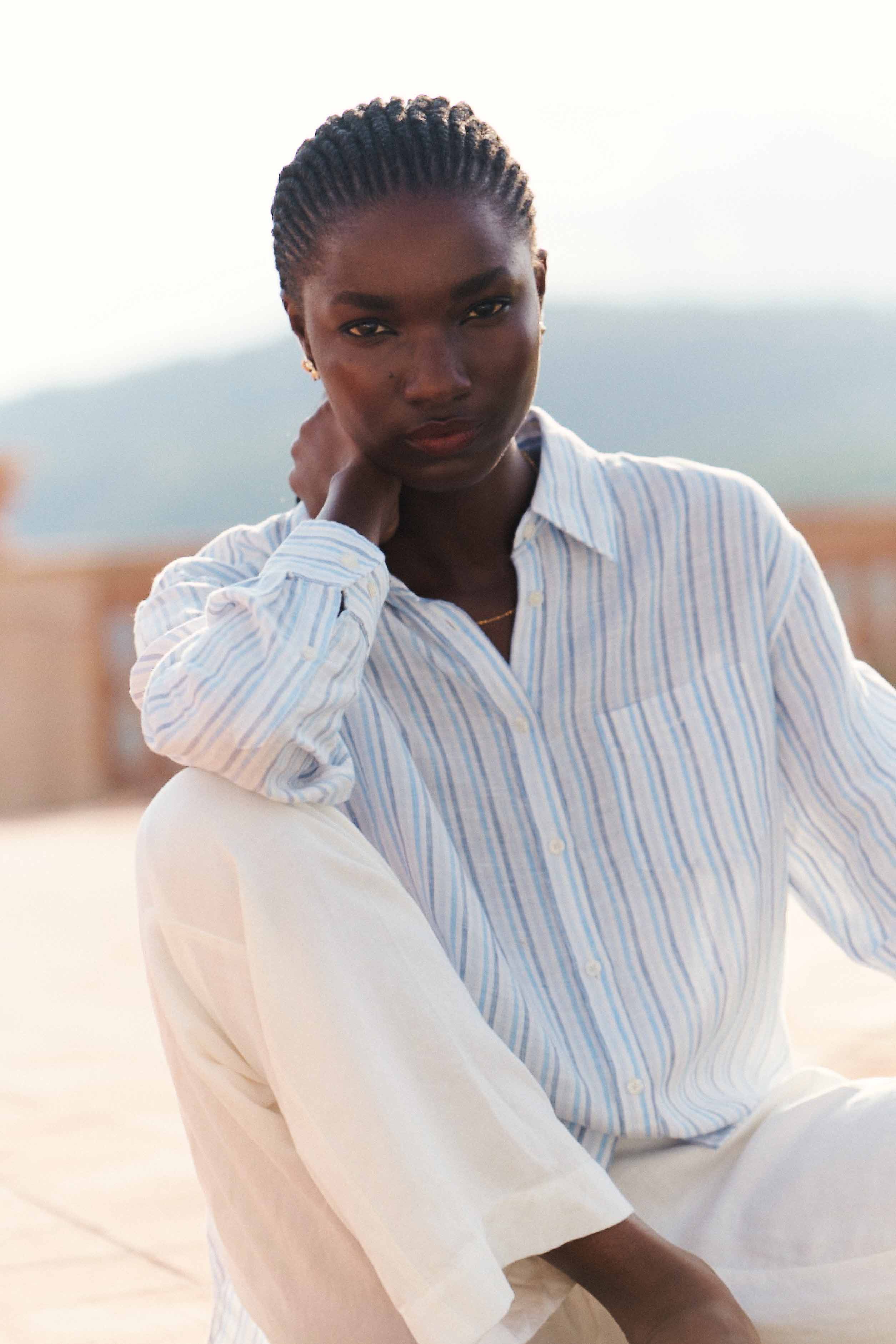 Women wearing a light, long‑sleeved striped shirt paired with relaxed white trousers, seated outdoors in a sunlit setting.