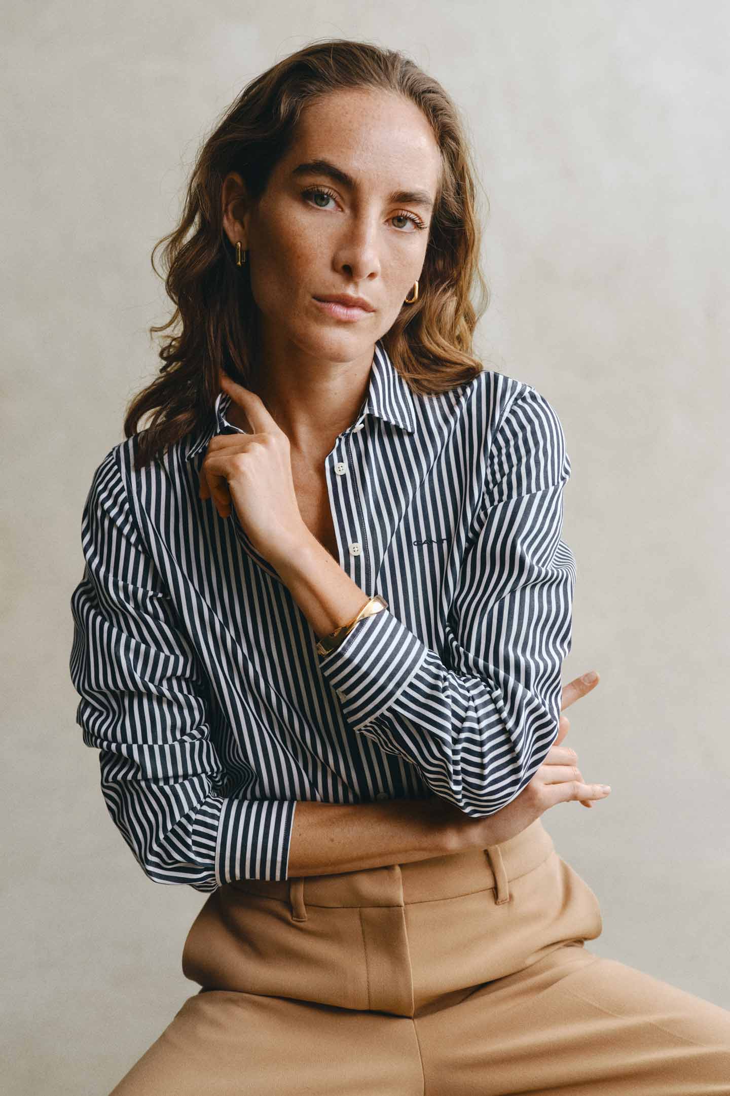 Model styled in a GANT striped shirt paired with beige pants seated in front of a grey backdrop.