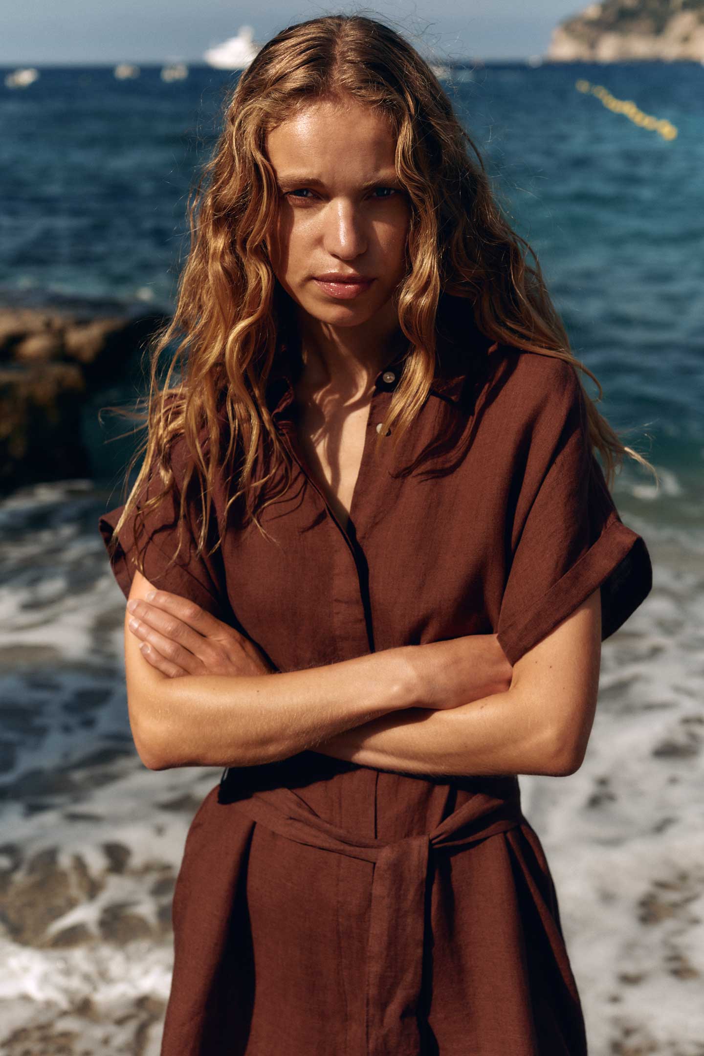 Woman wearing a Gant brown short-sleeve belted linen dress standing near the shoreline with the sea in the background.