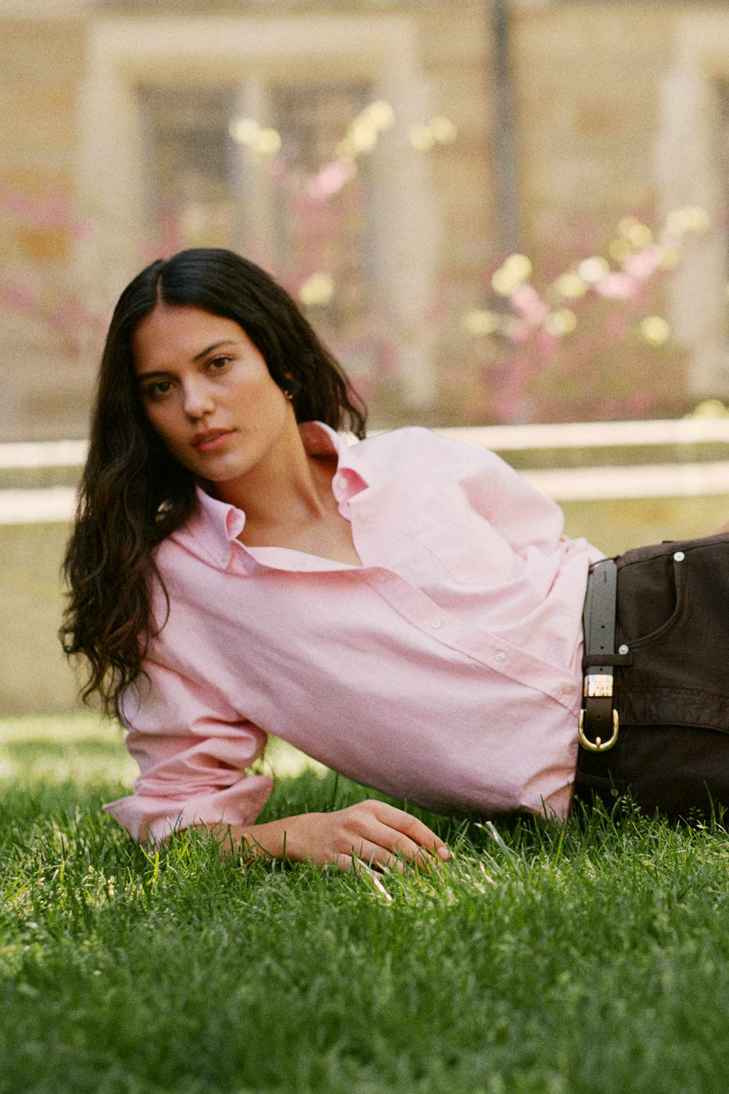 Model styled in a GANT pink shirt with dark jeans lying on grass in an outdoor campus setting.