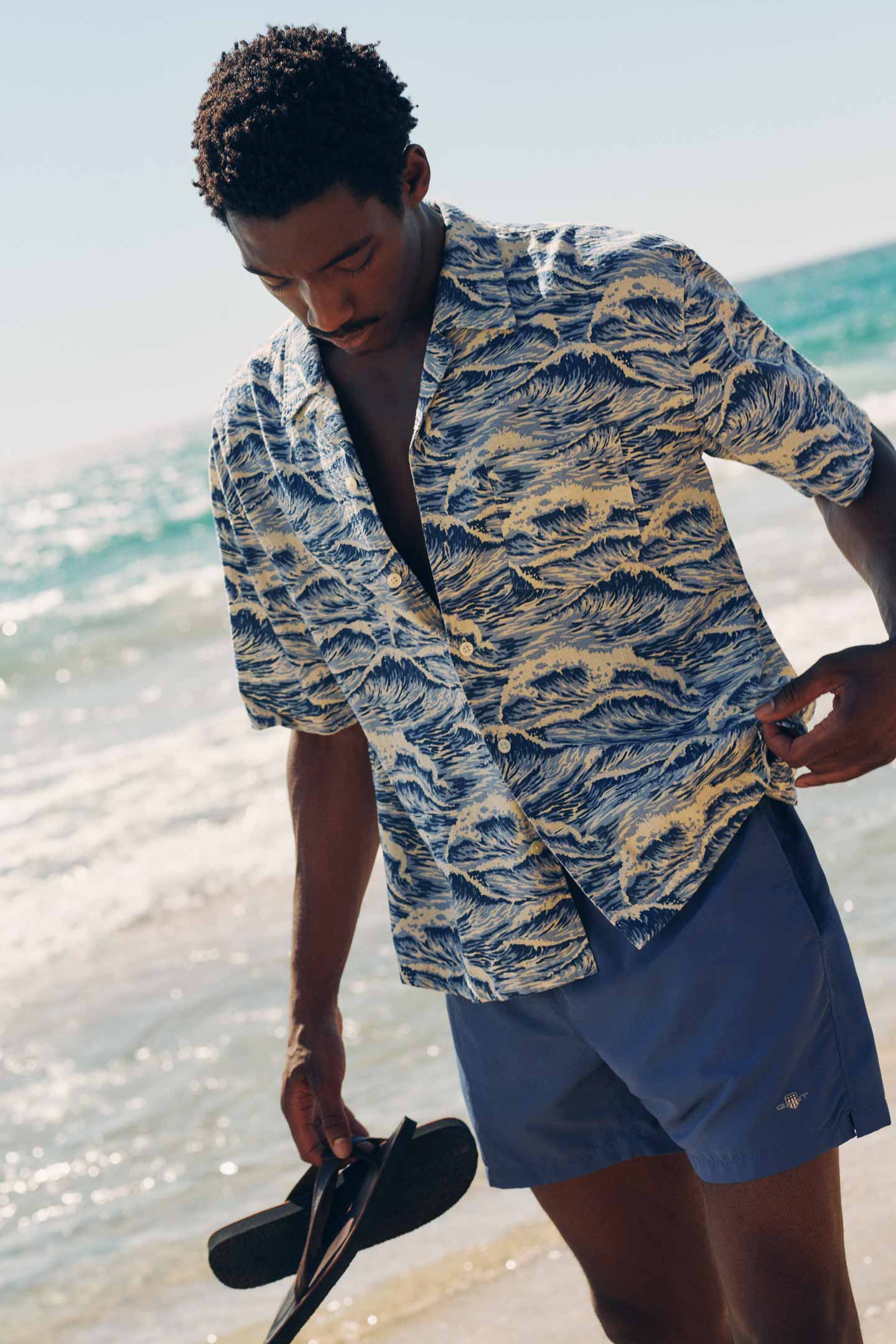 Man wearing a Gant printed short‑sleeve shirt and blue swim trunks while holding sandals on a sandy shoreline