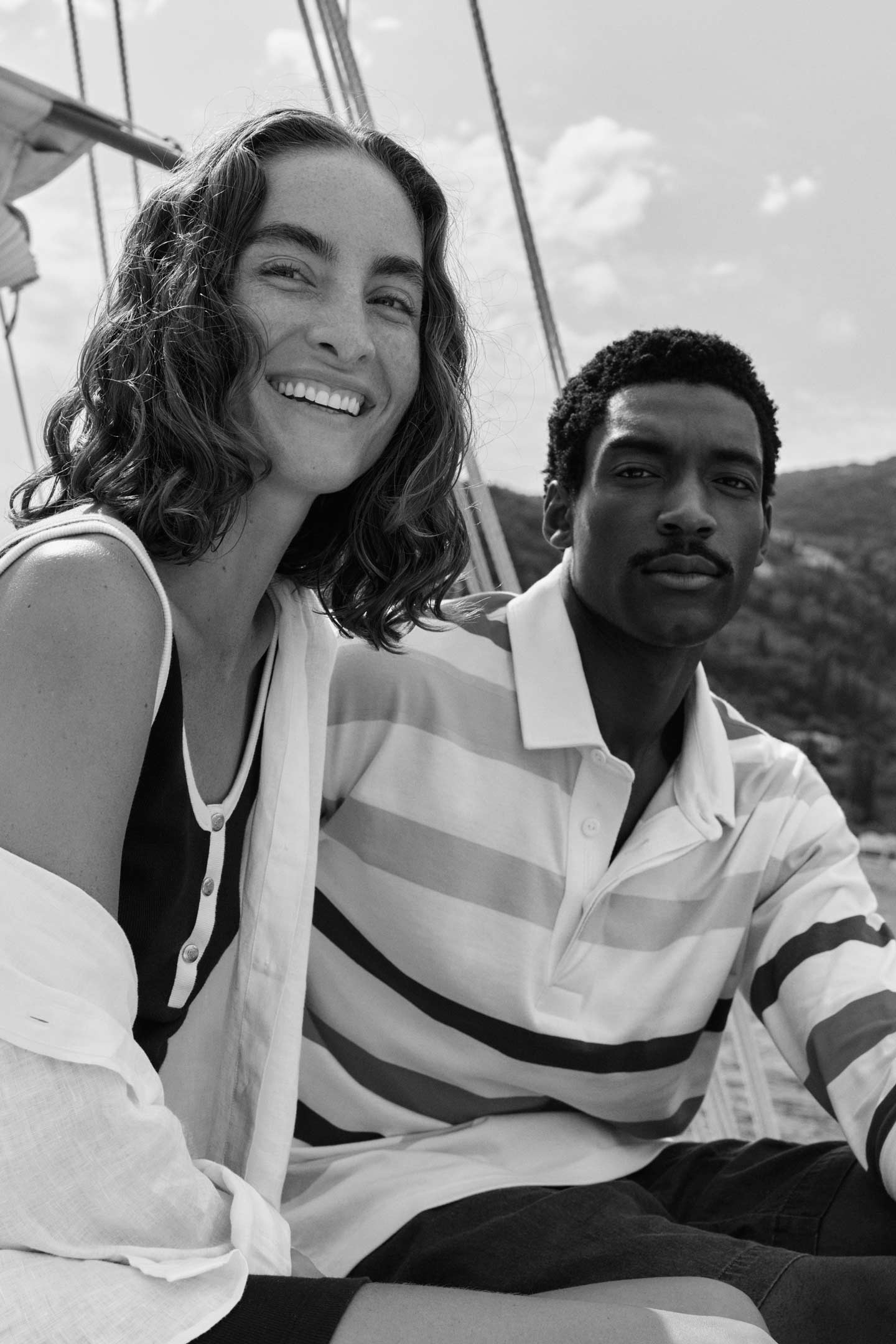 Couple on a boat, Man is wearing a multi striped Gant rugger and the Woman is wearing a sleeveless dress.