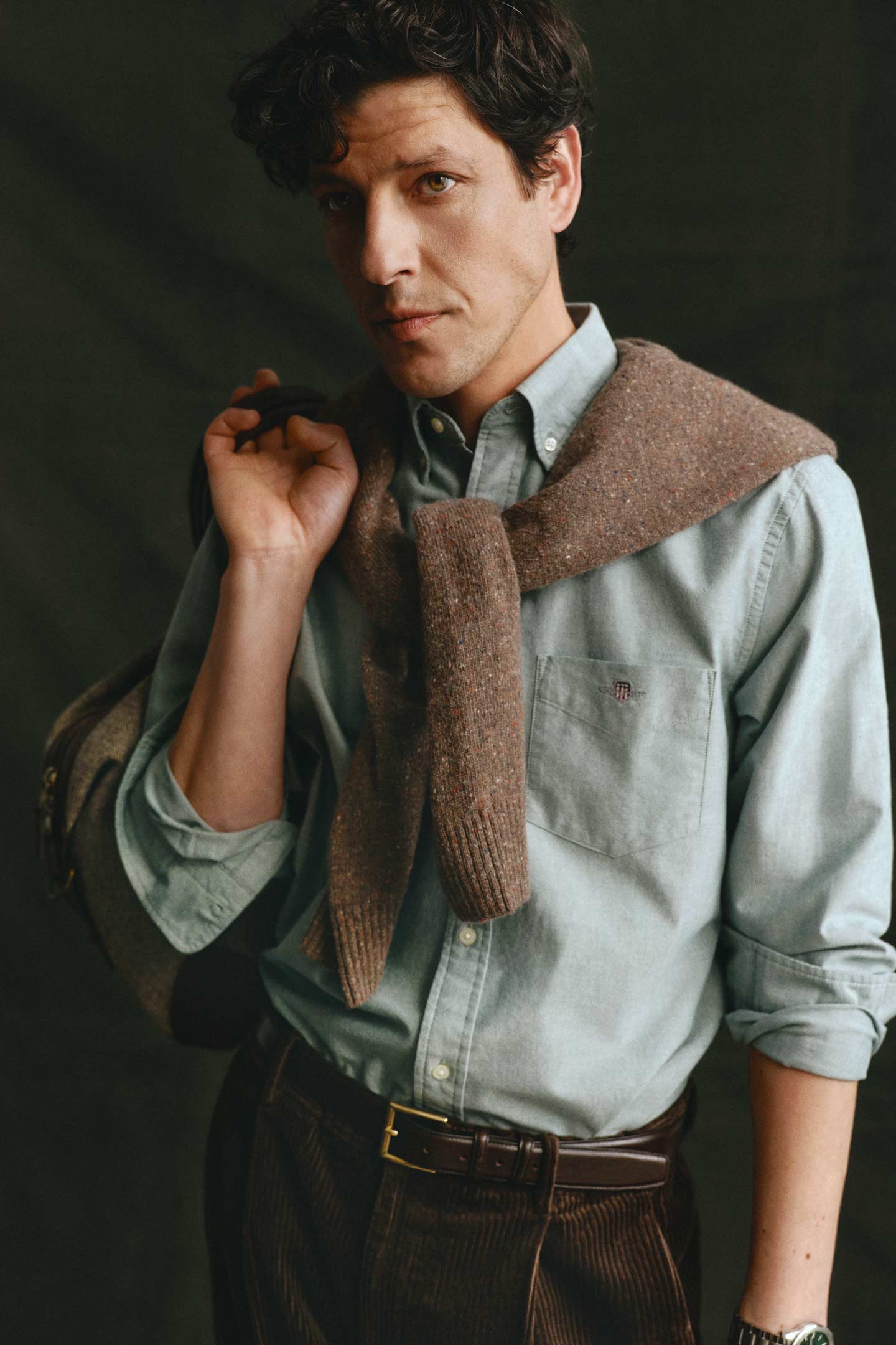 Model styled in a Gant button-down shirt styled with corduroy pants and a knit sweater draped over the shoulders.