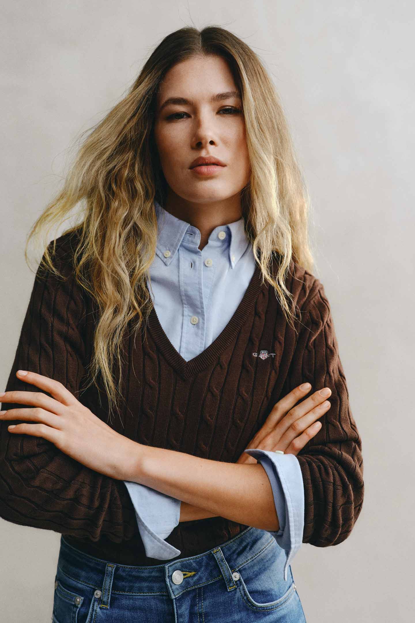 Model styled in a GANT brown cable v-neck knit layered over a blue shirt paired with blue denim jeans.