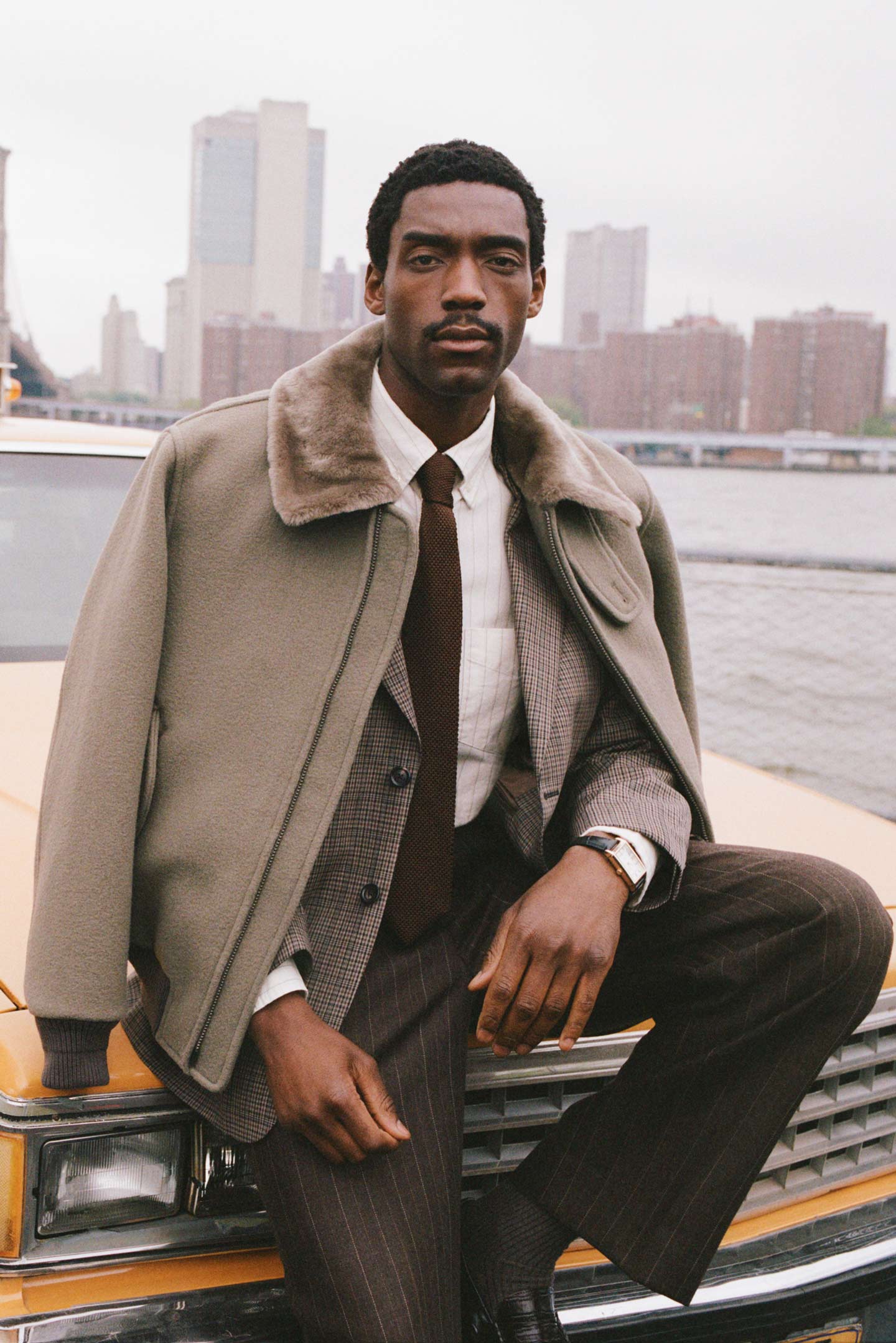 Model styled in a Gant bomber jacket with shearling collar over a checked blazer, shirt and tie, seated on a yellow taxi.