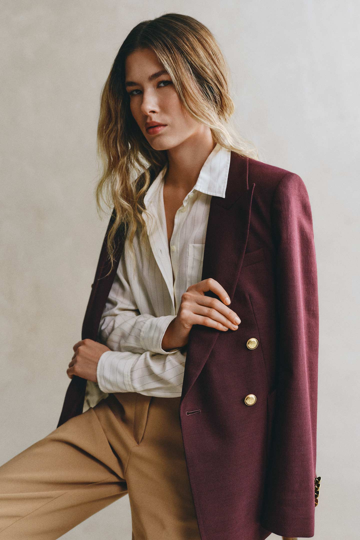 Model styled in a GANT white striped shirt with dark khaki pants with a dark red blazer over shoulders sitting on a chair in front of a grey backdrop.