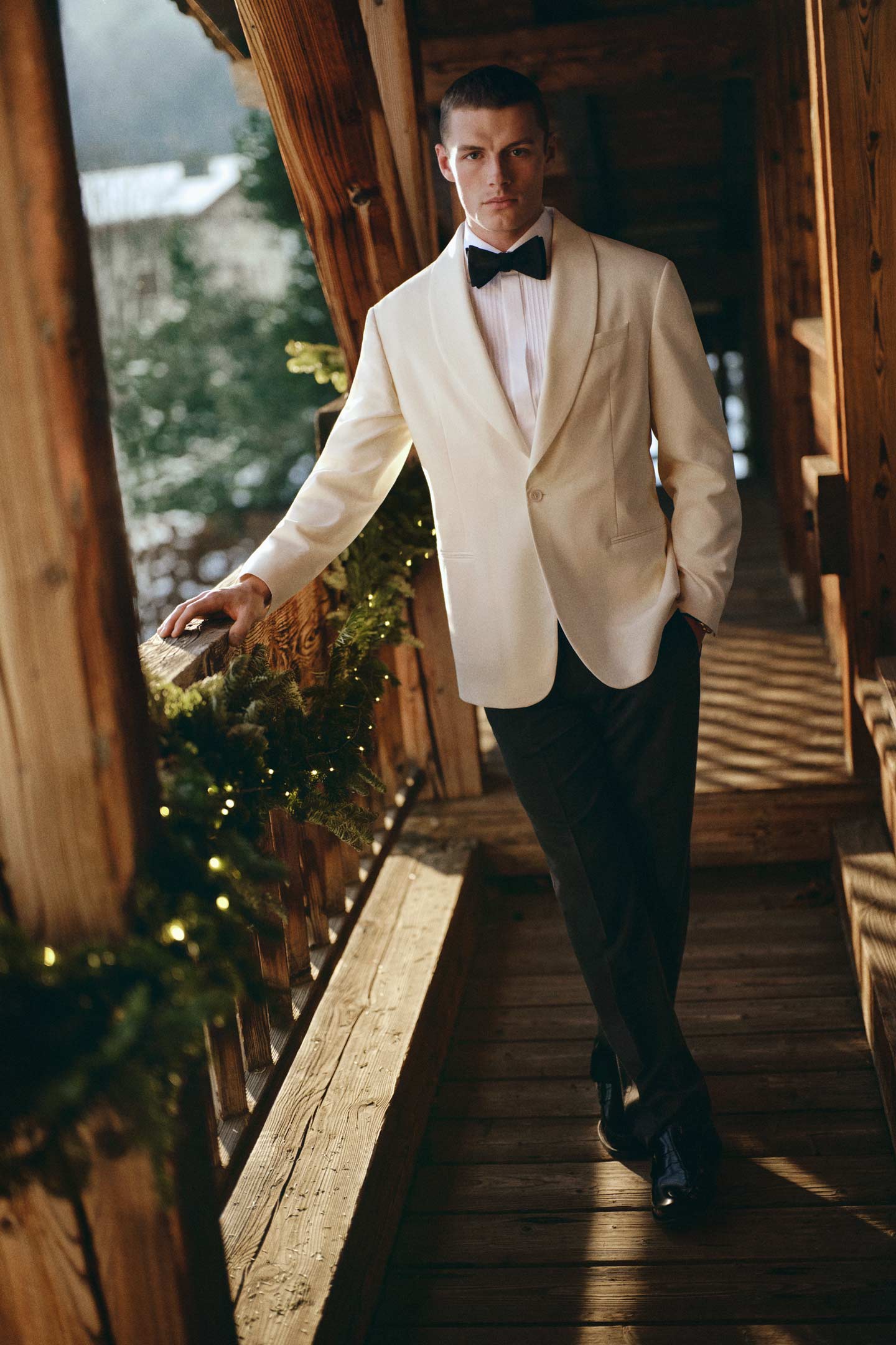 Model styled in a white Gant tuxedo jacket paired with black pants and a bow tie, standing on a wooden lodge balcony.