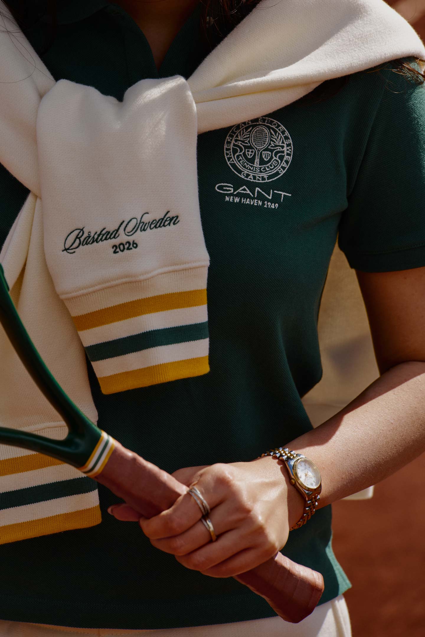 Woman wearing a Gant green polo shirt with a sweater tied over the shoulders while holding a tennis racket in an outdoor setting.