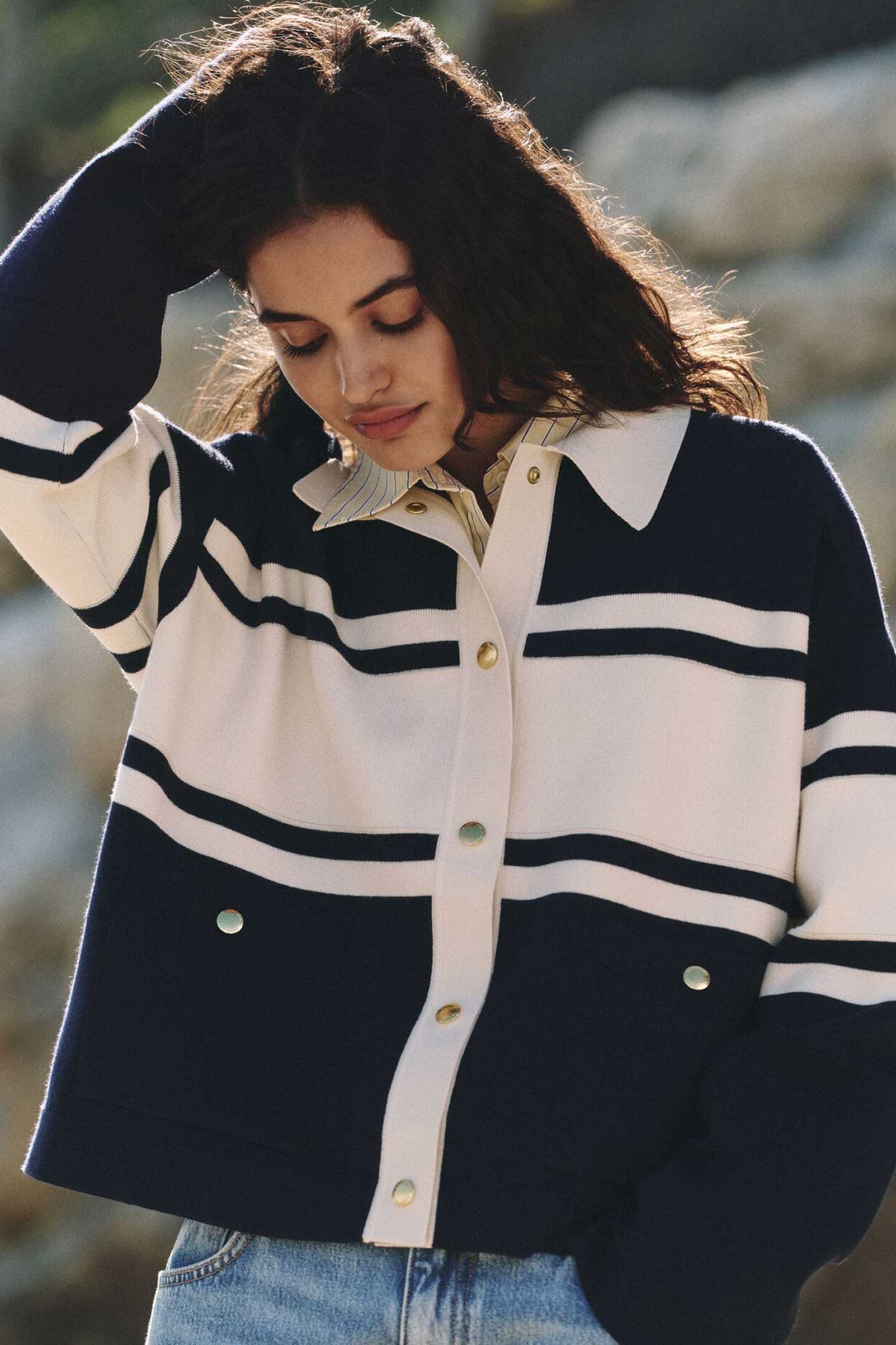 Woman wearing a Gant navy and cream striped cardigan with gold buttons over a collared shirt, standing outdoors against a rocky backdrop. One arm is raised to adjust her hair, highlighting the casual, coastal-inspired style.