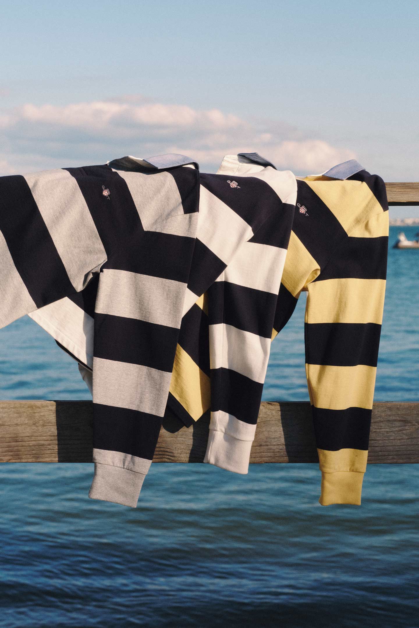 Gant striped rugby shirts in blue‑grey, blue‑white, and blue‑yellow draped over a wooden railing by the water.