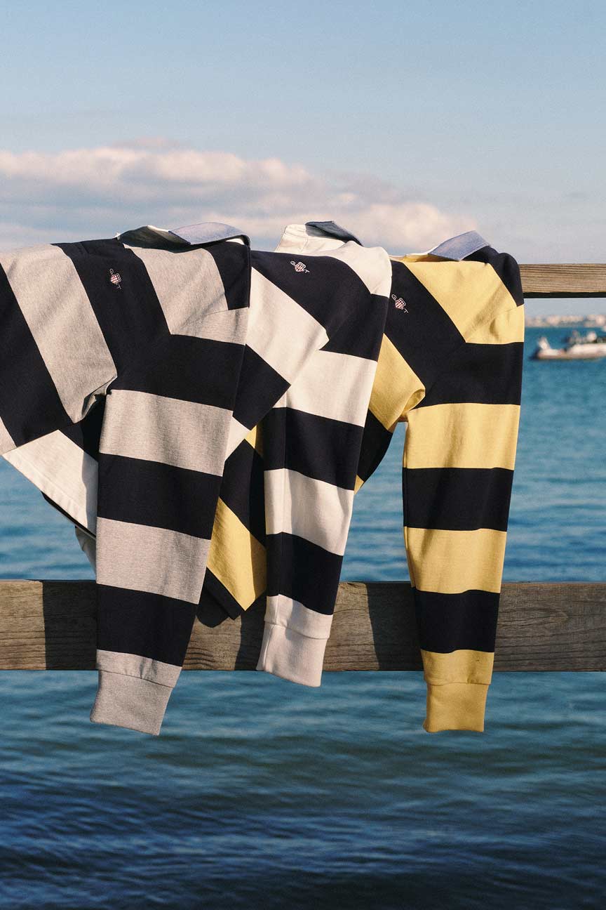 Gant striped rugby shirts in blue‑grey, blue‑white, and blue‑yellow draped over a wooden railing by the water.