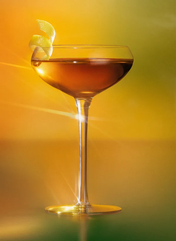 Lagavulin Sweet Peat Tea cocktail served in a coupe glass with lemon twist, showcasing the whisky's golden amber color against a warm gradient background.