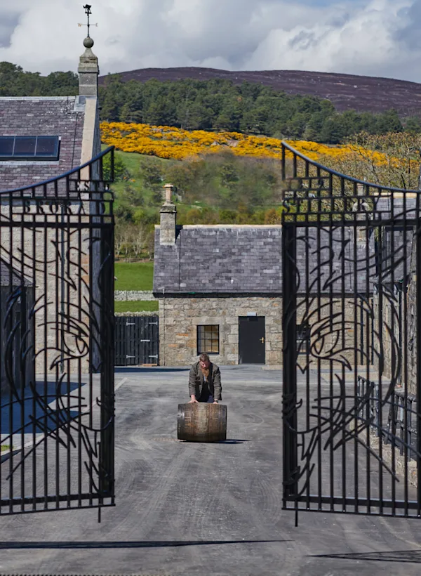 A person rolls a wooden barrel towards the gates of Brora distillery, which are slightly open. Behind the person can be seen some of Brora’s buildings, and behind those are tall hills with assorted trees and grasses.