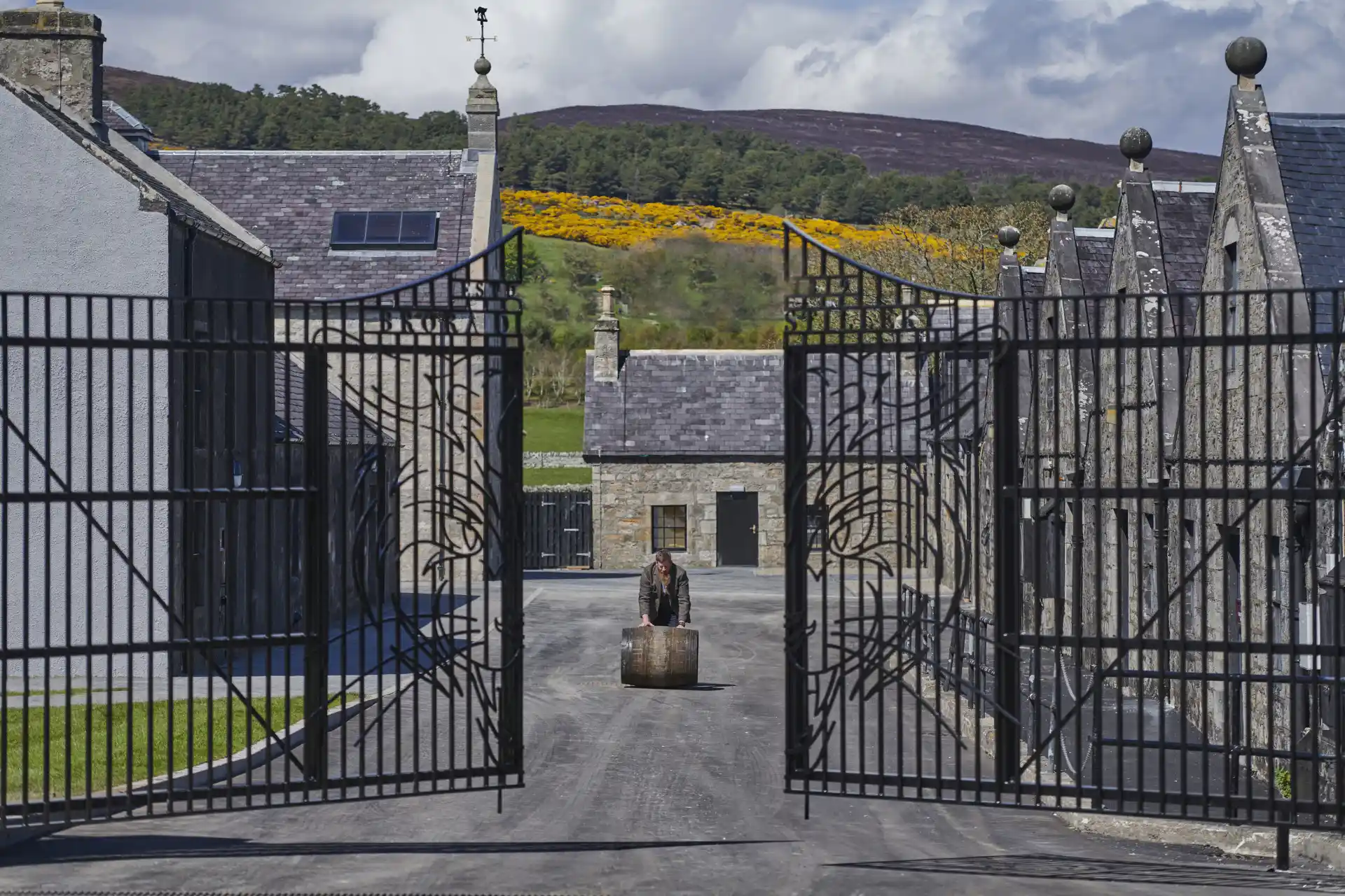 A person rolls a wooden barrel towards the gates of Brora distillery, which are slightly open. Behind the person can be seen some of Brora’s buildings, and behind those are tall hills with assorted trees and grasses.