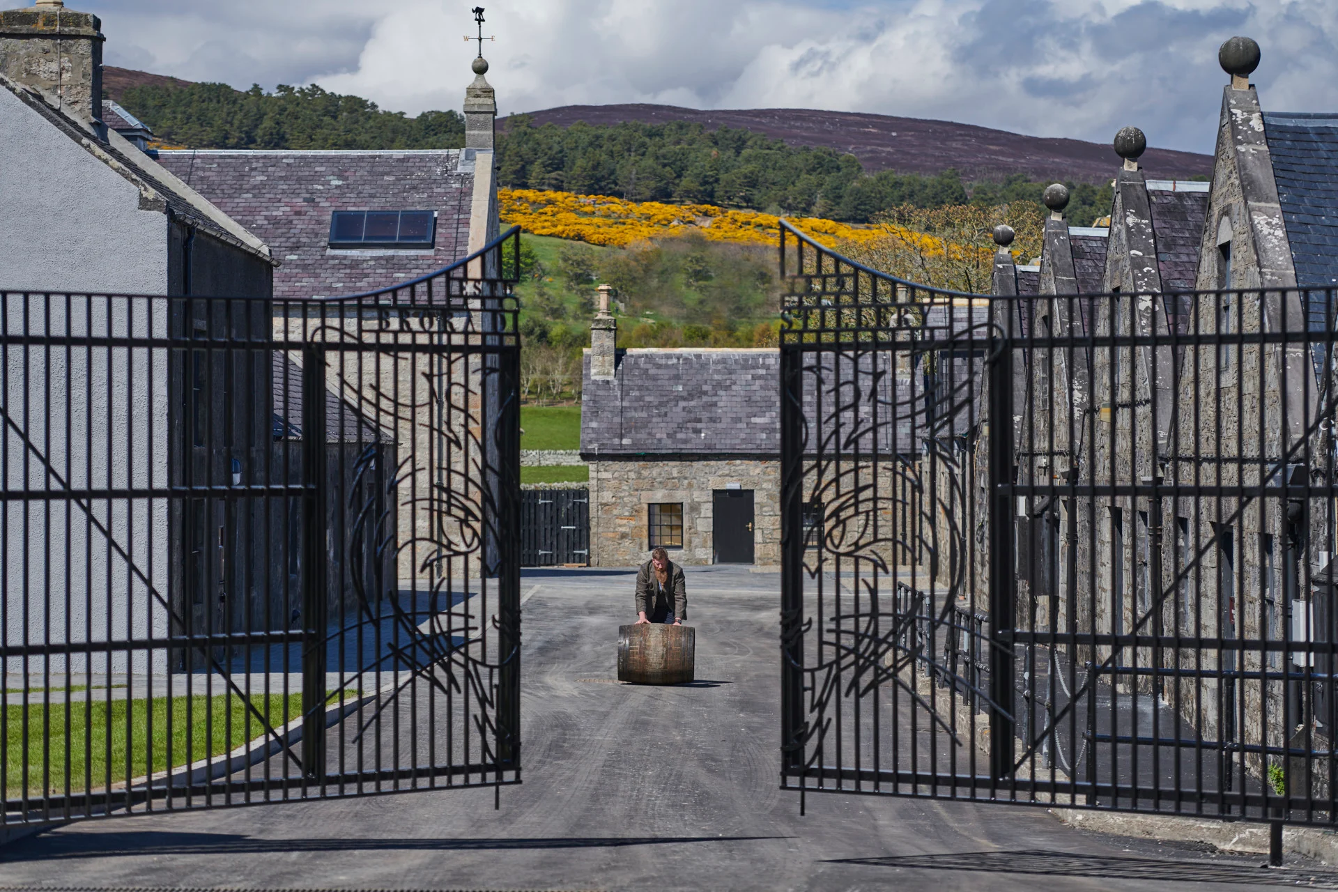 A person rolls a wooden barrel towards the gates of Brora distillery, which are slightly open. Behind the person can be seen some of Brora’s buildings, and behind those are tall hills with assorted trees and grasses.