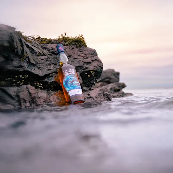 Een fles Talisker The Wild Blue staat op de rotsachtige kustlijn terwijl de zee op de achtergrond spuit