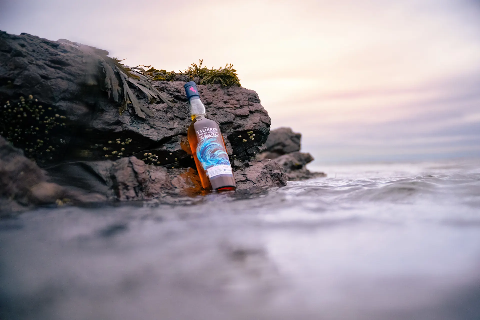 Een fles Talisker The Wild Blue staat op de rotsachtige kustlijn terwijl de zee op de achtergrond spuit